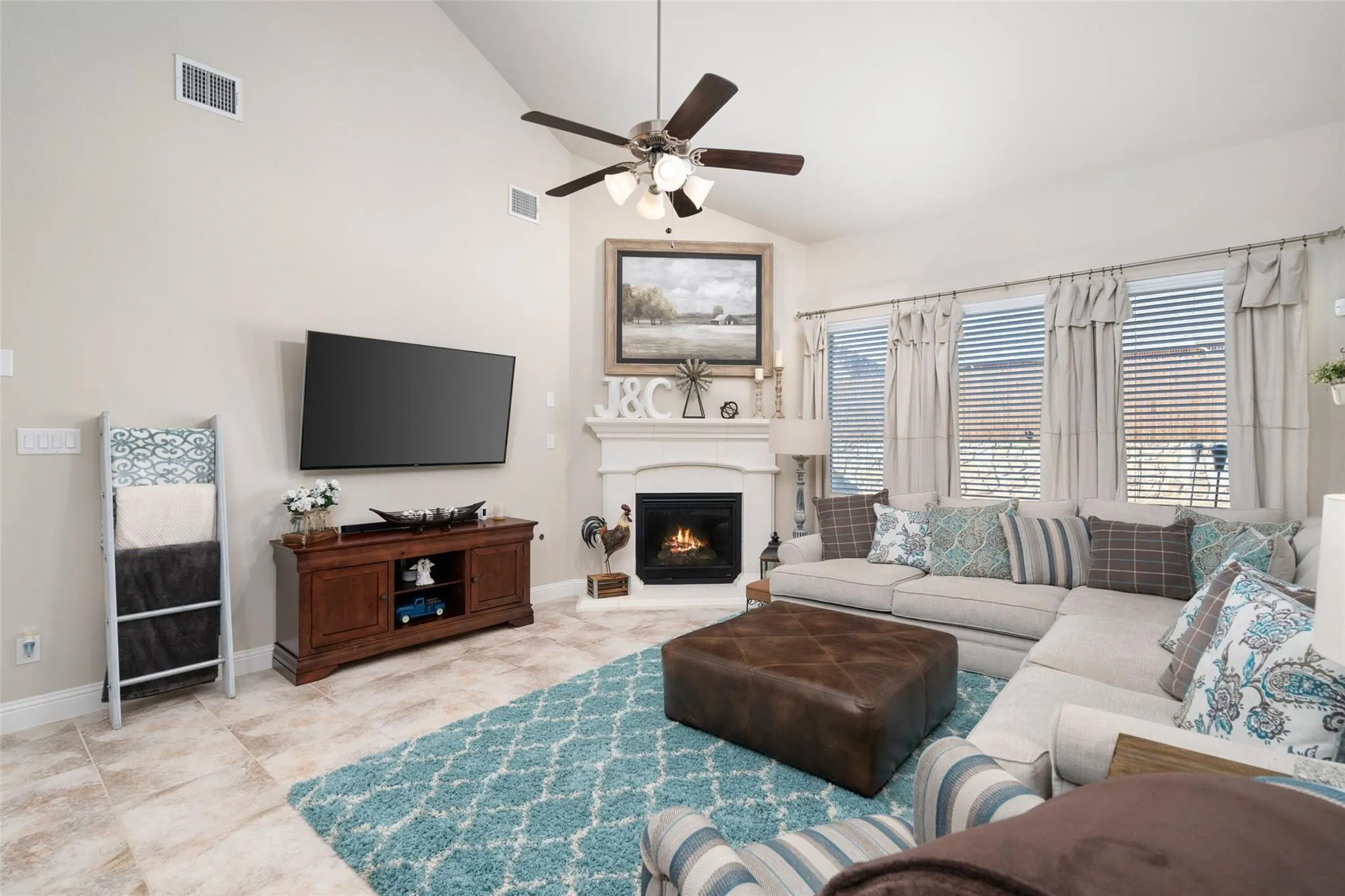 Vaulted ceiling with a ceiling fan, corner fireplace with a mantel, multiple windows with blinds, and tile flooring