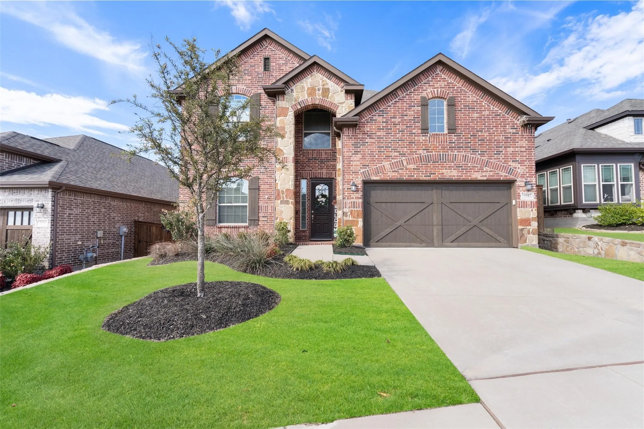 Brick and stone facade, arched entryway, dark wood garage doors, concrete driveway, and manicured landscaping