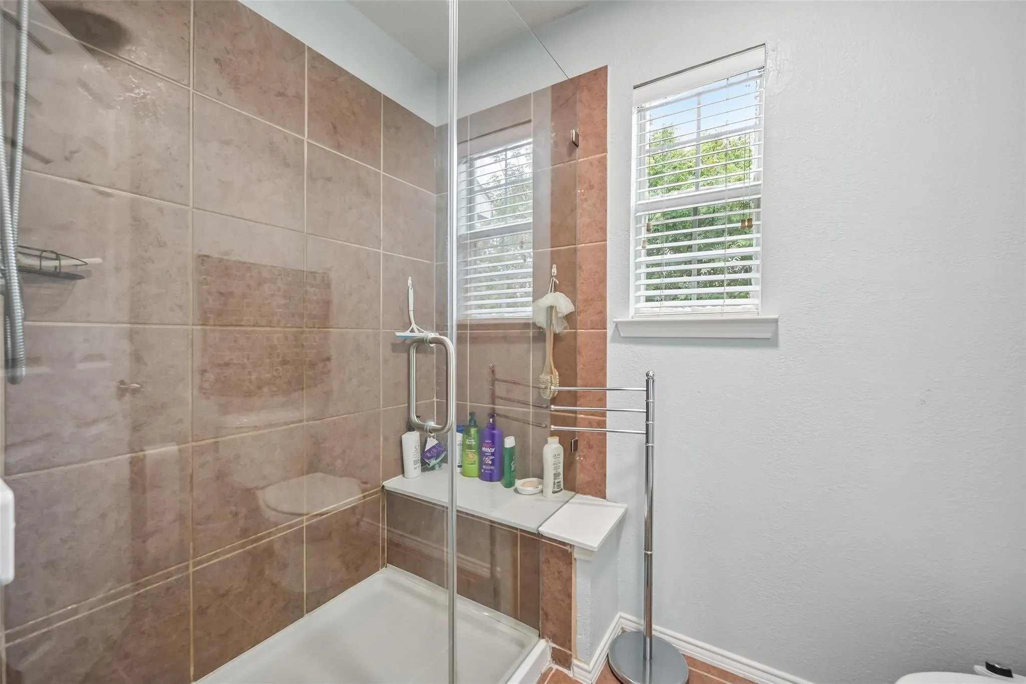 Glass-enclosed shower with a built-in bench, brown tile surround, and dual windows with blinds