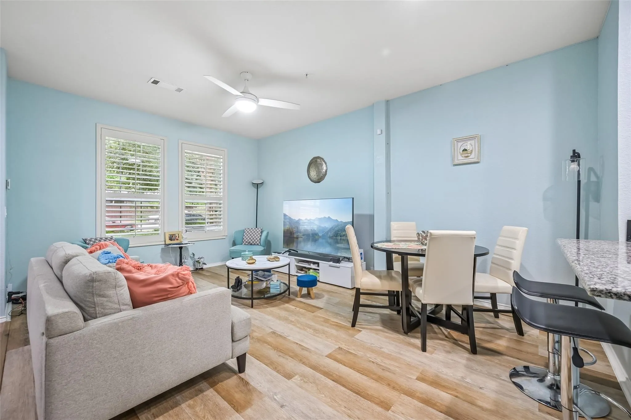 Wood-finish flooring throughout, two large windows with plantation shutters, a ceiling fan with integrated lighting, a built-in breakfast bar with a stone-finish countertop, and a dining area