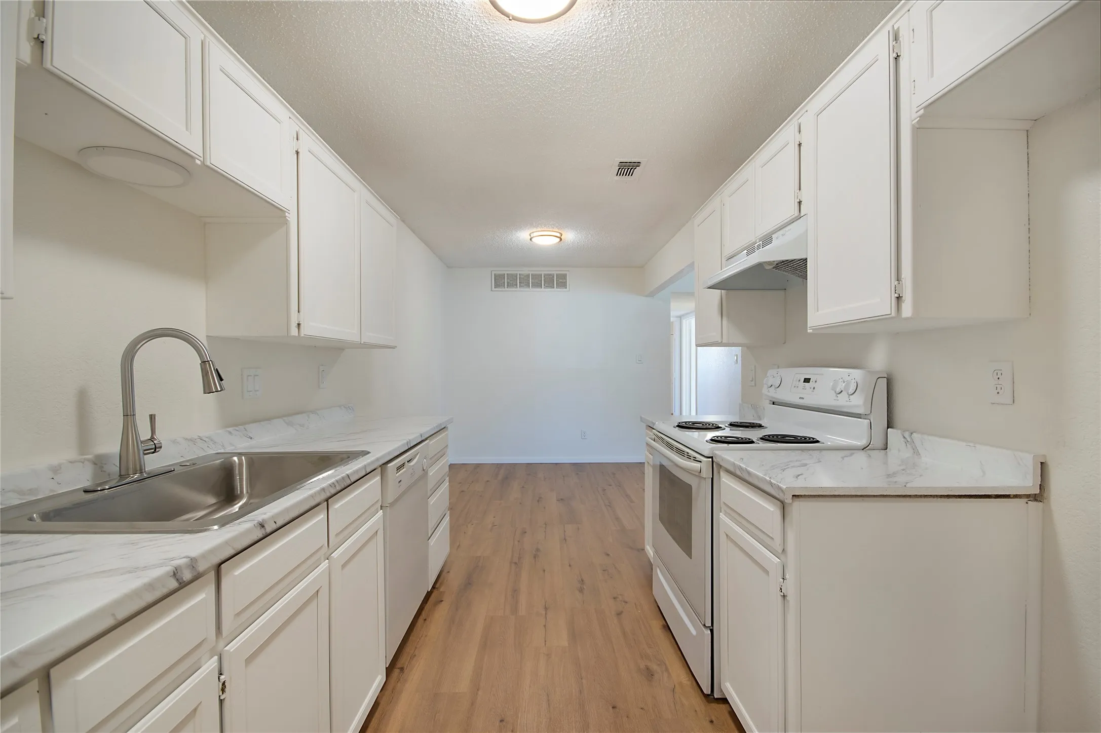 Galley kitchen featuring wood-finish flooring, white cabinetry, a double basin stainless steel sink, and white appliances