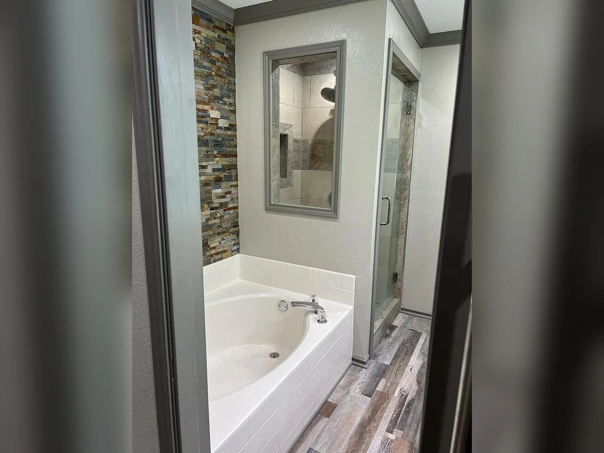 Corner soaking tub, stone-finish accent wall, glass-enclosed shower, wood-finish flooring, and crown molding