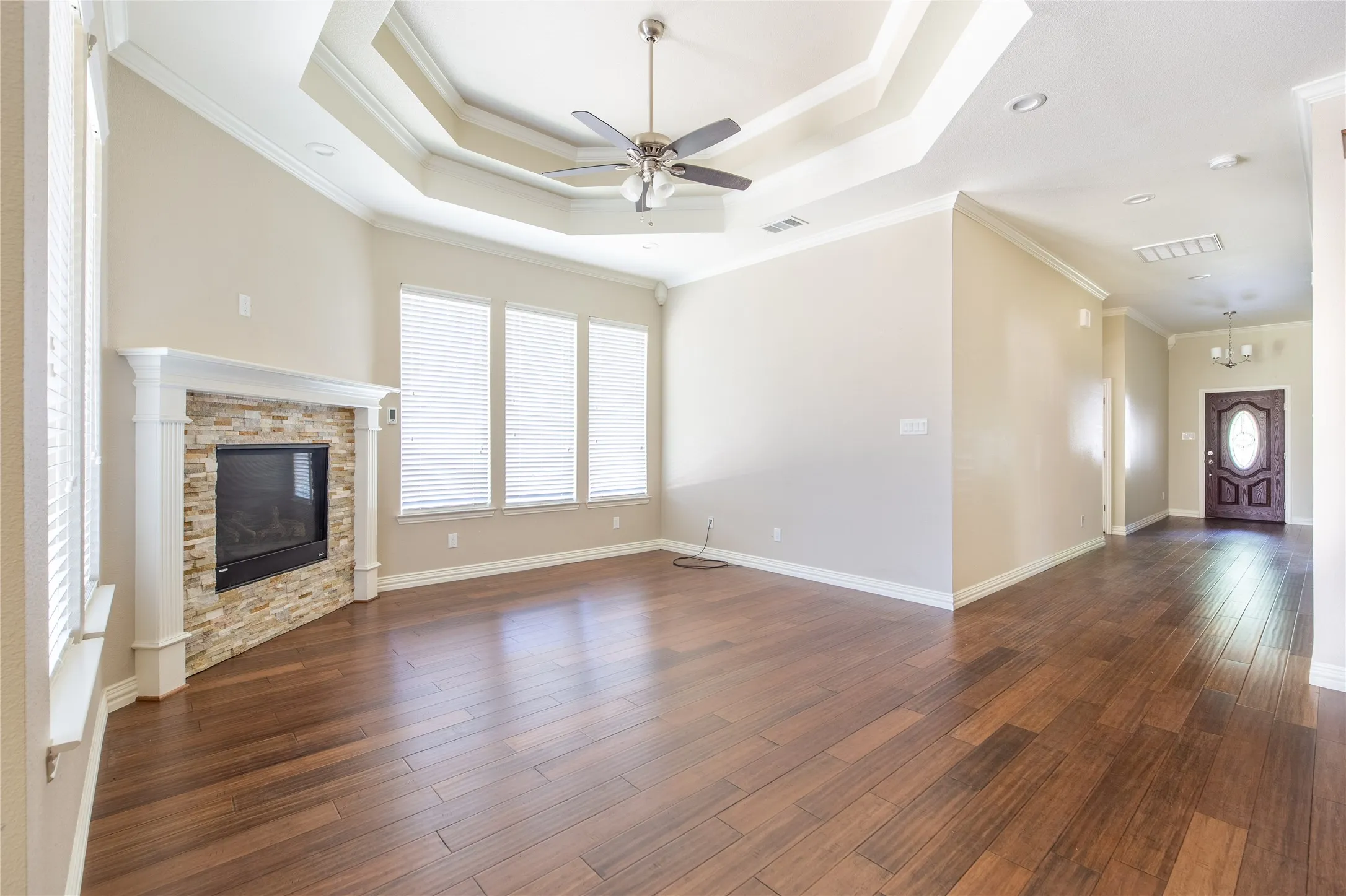 Spacious living area featuring wood-finish flooring, a tray ceiling with a ceiling fan, a fireplace with a stacked stone facade, and multiple windows