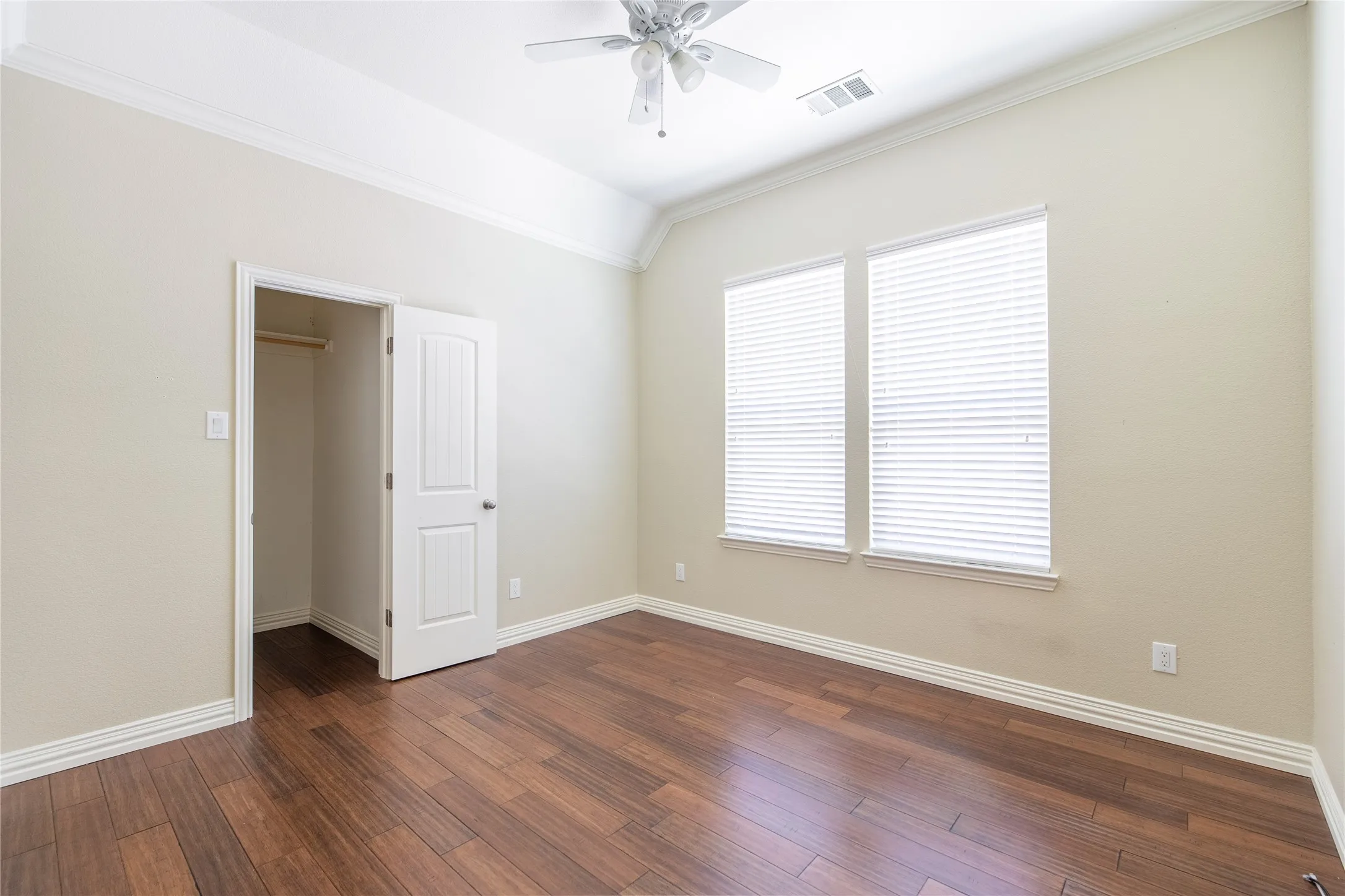 Vaulted ceiling with crown molding, wood-finish flooring, two large windows with blinds, and a ceiling fan with light fixture