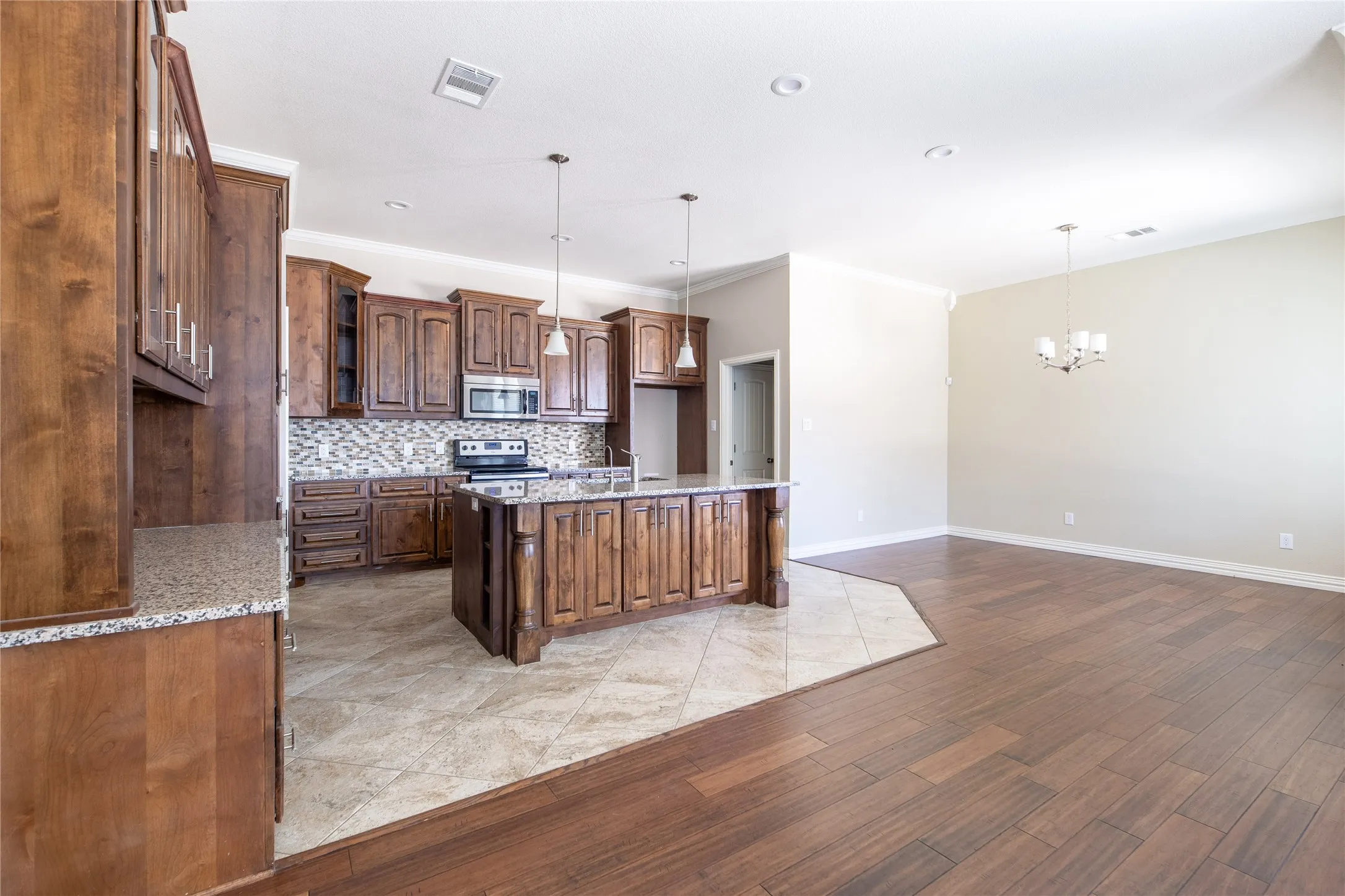 Open-concept kitchen and living area featuring rich wood cabinetry with decorative hardware, stainless steel appliances, mosaic tile backsplash, granite countertops, a substantial island with seating, recessed lighting, and warm wood-finish flooring throughout the main living space