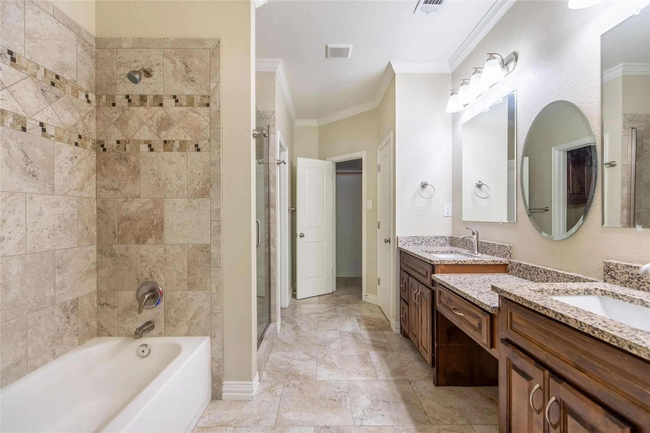 Spa-inspired primary bathroom featuring dual vanities with granite countertops, an oval mirror with integrated lighting, a walk-in shower with travertine tile surround and multiple body jets, and travertine flooring throughout