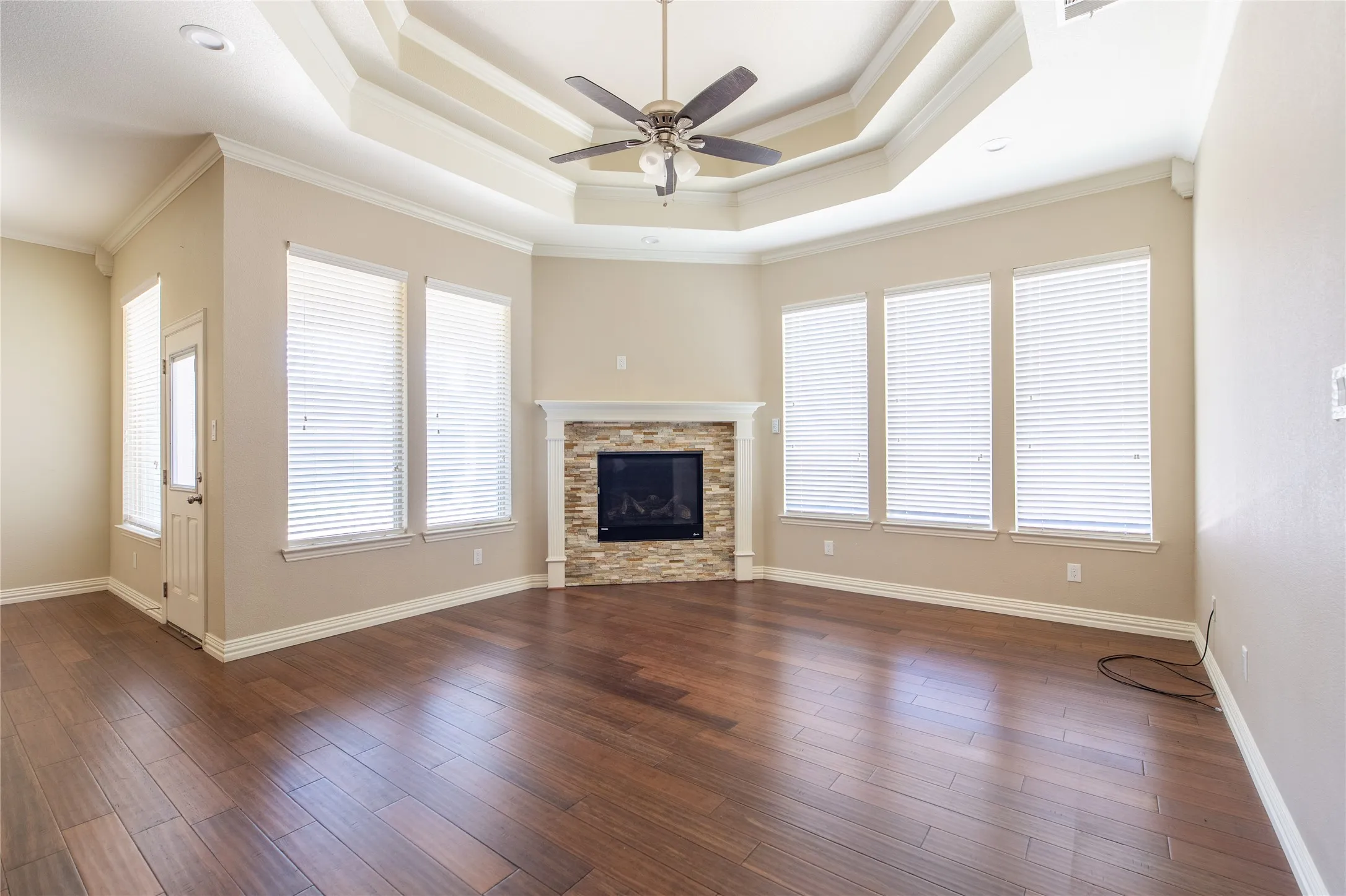 Expansive living area featuring a tray ceiling, wood-finish flooring, a corner fireplace with a stone surround, and multiple windows