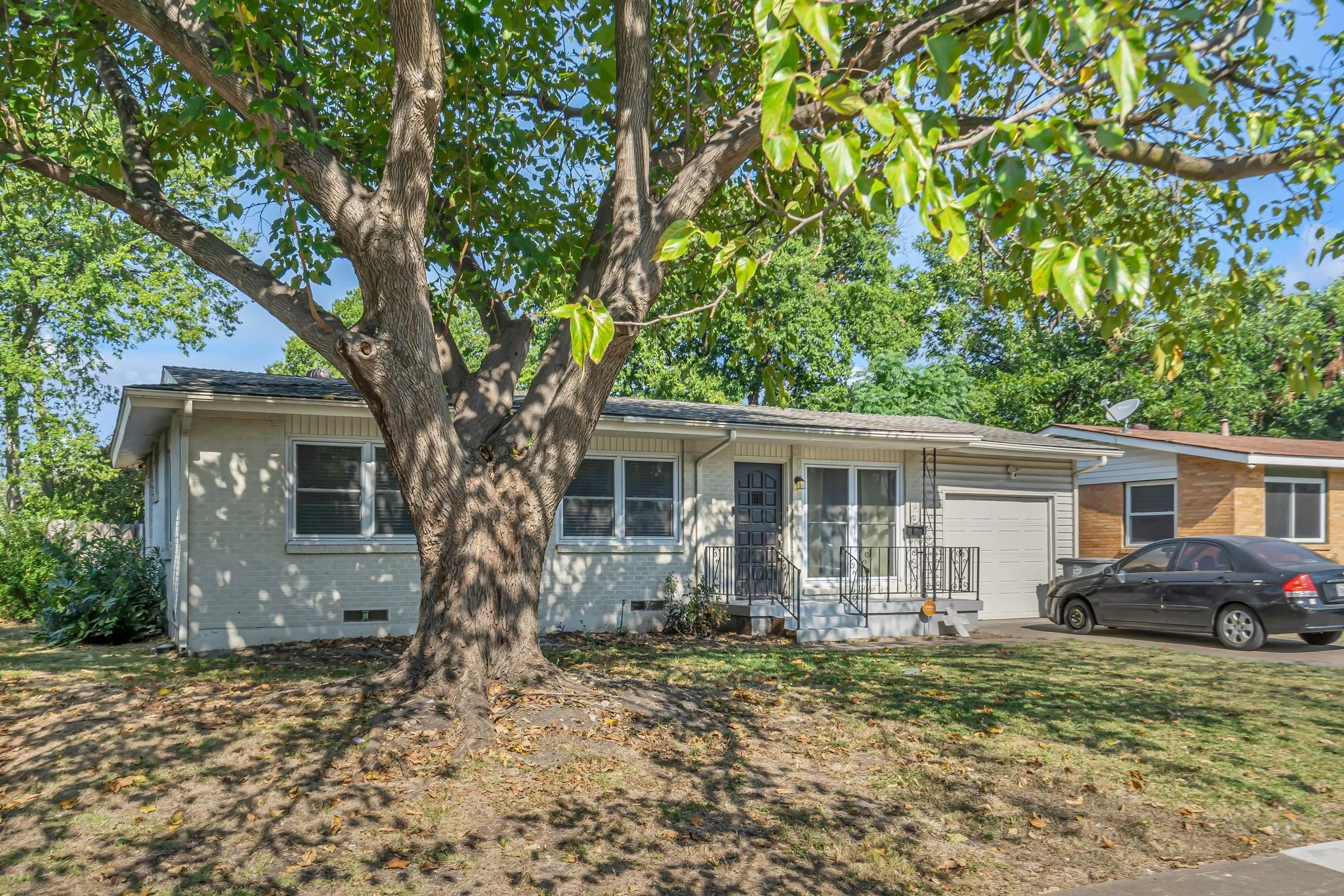 Exterior featuring a large mature tree, light-colored stucco siding, a covered front entry with railing, and an attached garage with a corrugated door