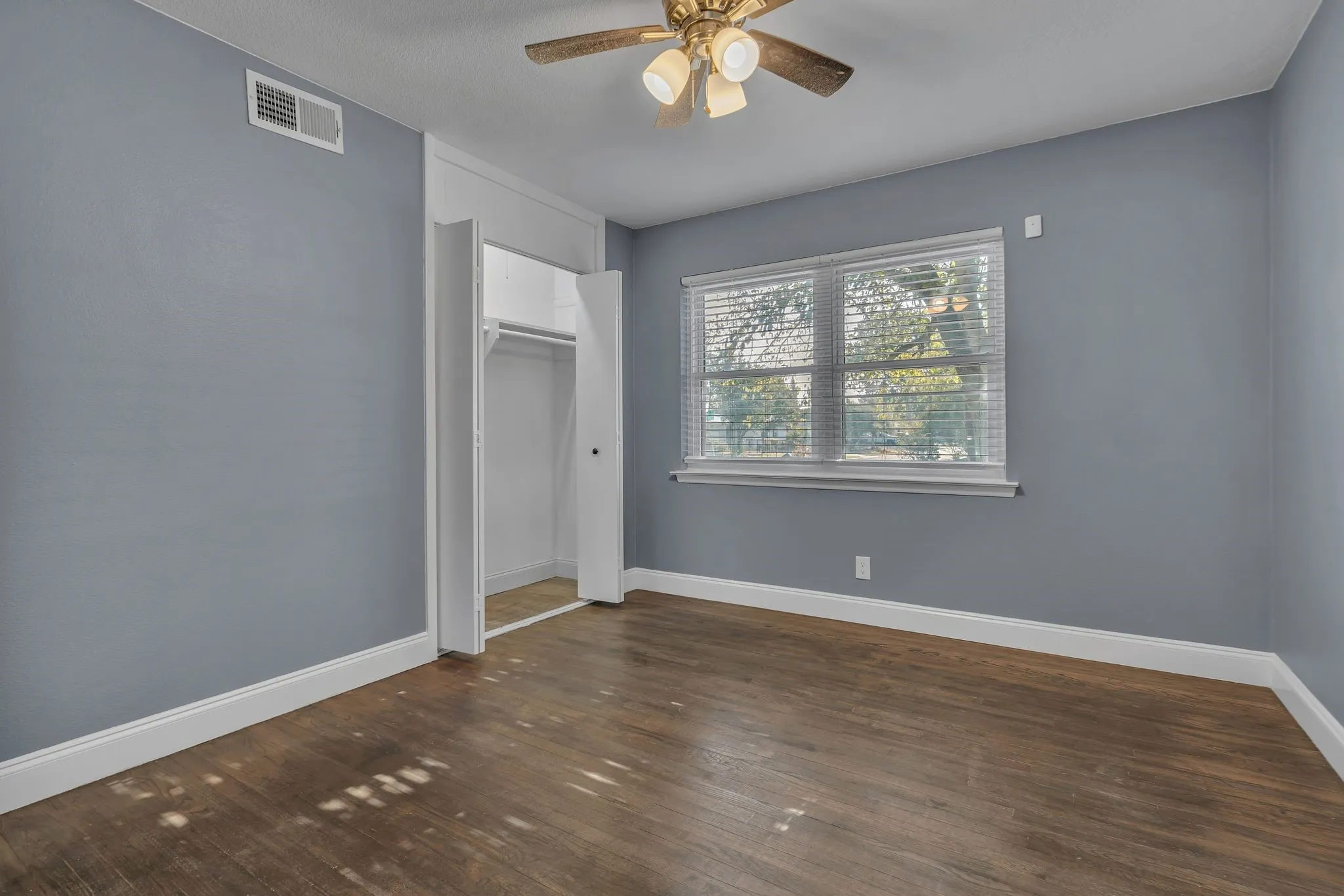 Wood-finish flooring, light blue wall paint, a ceiling fan with integrated lighting, a window with horizontal blinds, and a built-in closet with bi-fold doors