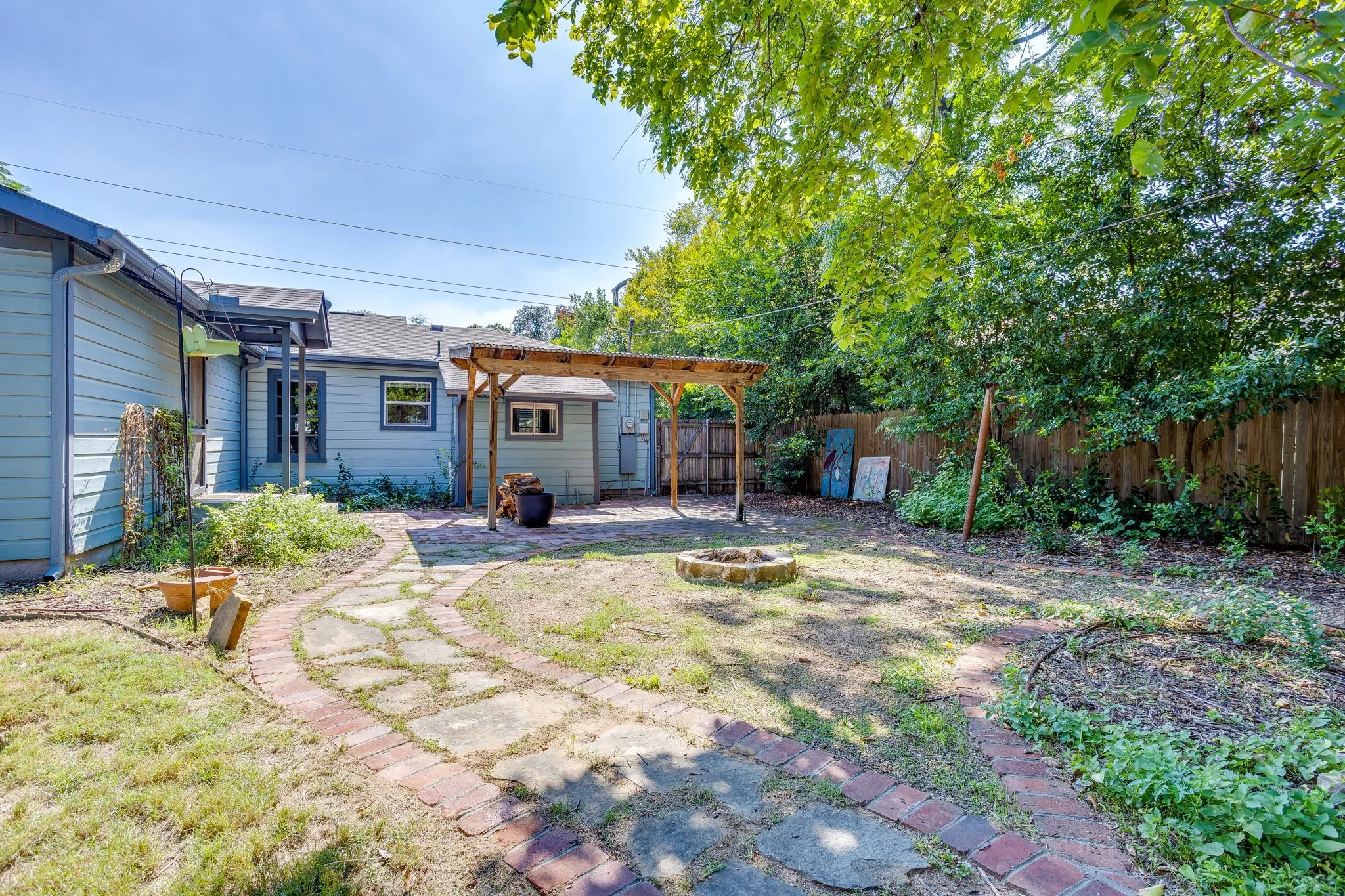 Expansive backyard featuring a stone and brick pathway, a fire pit, mature trees, and a wooden pergola covering a paved patio