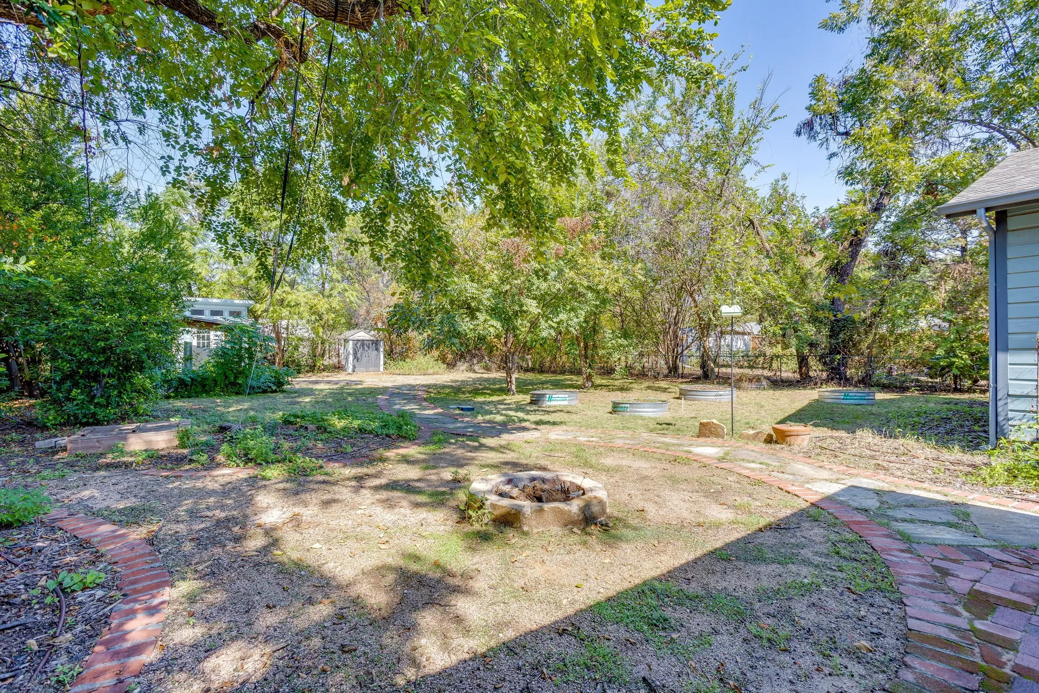 Expansive backyard featuring mature trees, a stone-lined fire pit, and a brick paver pathway