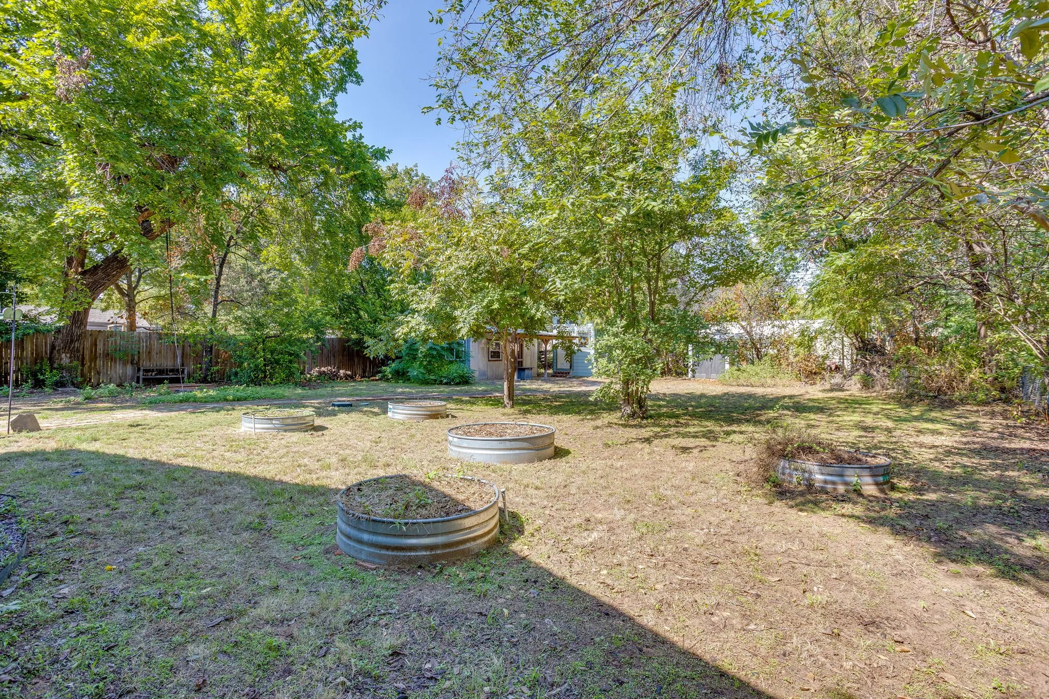 Spacious backyard featuring multiple mature trees, a wooden privacy fence, and several metal raised garden beds