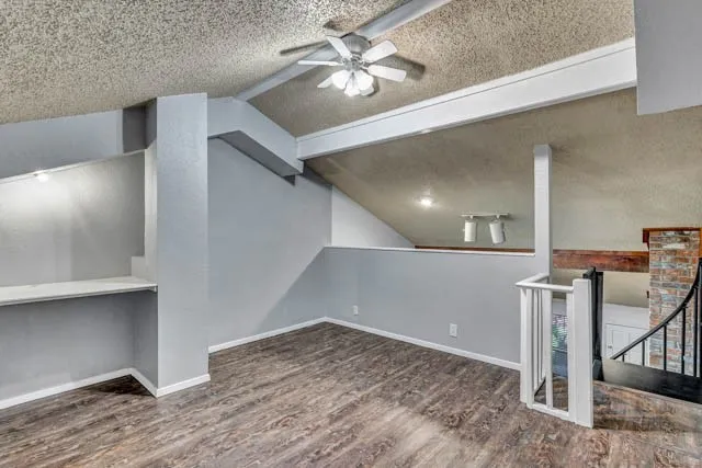 Vaulted ceilings with exposed structural beams, wood-finish flooring, built-in shelving, white balustrade railing, and brick accent feature