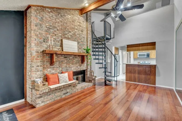 Full-height brick fireplace featuring a wood mantel, vaulted ceiling with exposed wood beam, spiral staircase with metal railing, and wood-finish flooring