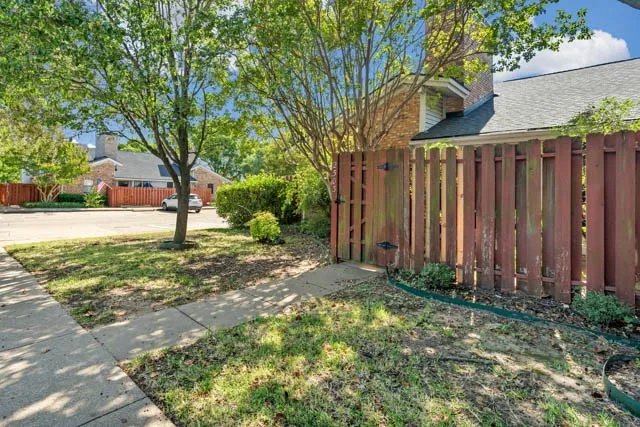 Wood-slat privacy fencing with gated access, concrete sidewalk, masonry chimney, and mature shade trees