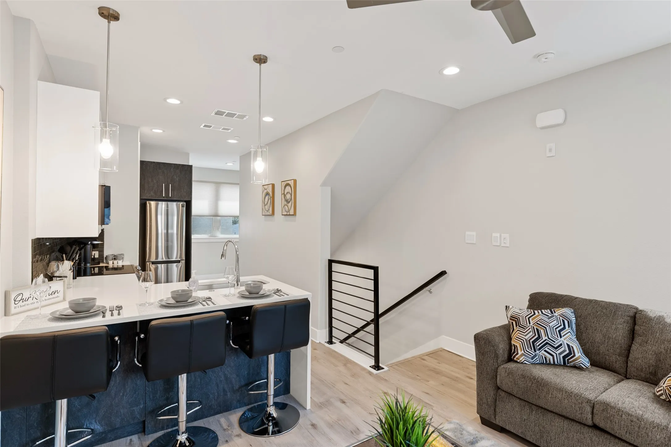 Kitchen island with a white countertop and seating, stainless steel appliances, modern pendant lighting, wood-finish flooring, and a contemporary staircase with metal railings