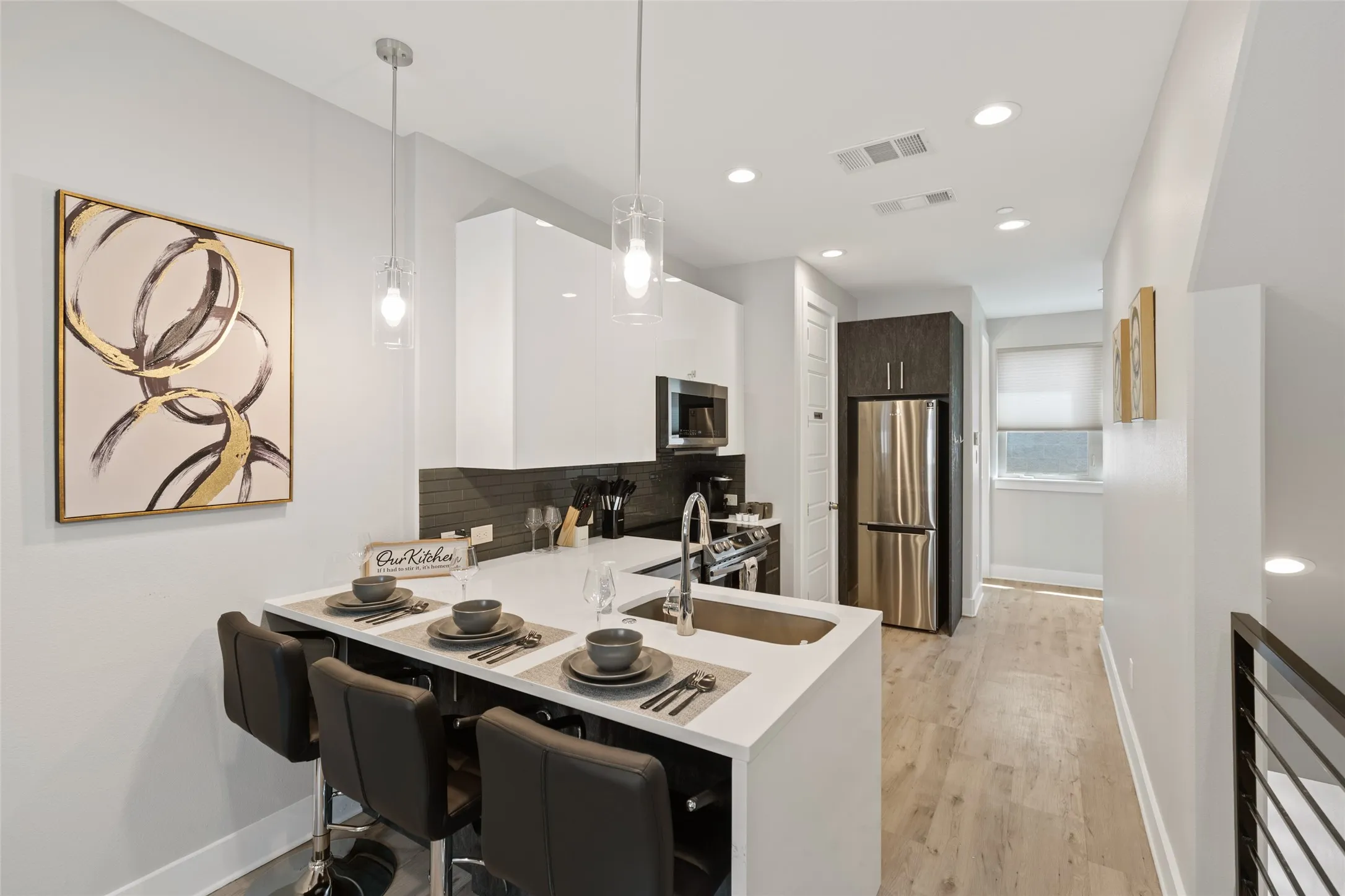 Modern kitchen featuring a white countertop island with integrated sink, stainless steel appliances, dark tile backsplash, and wood-finish flooring