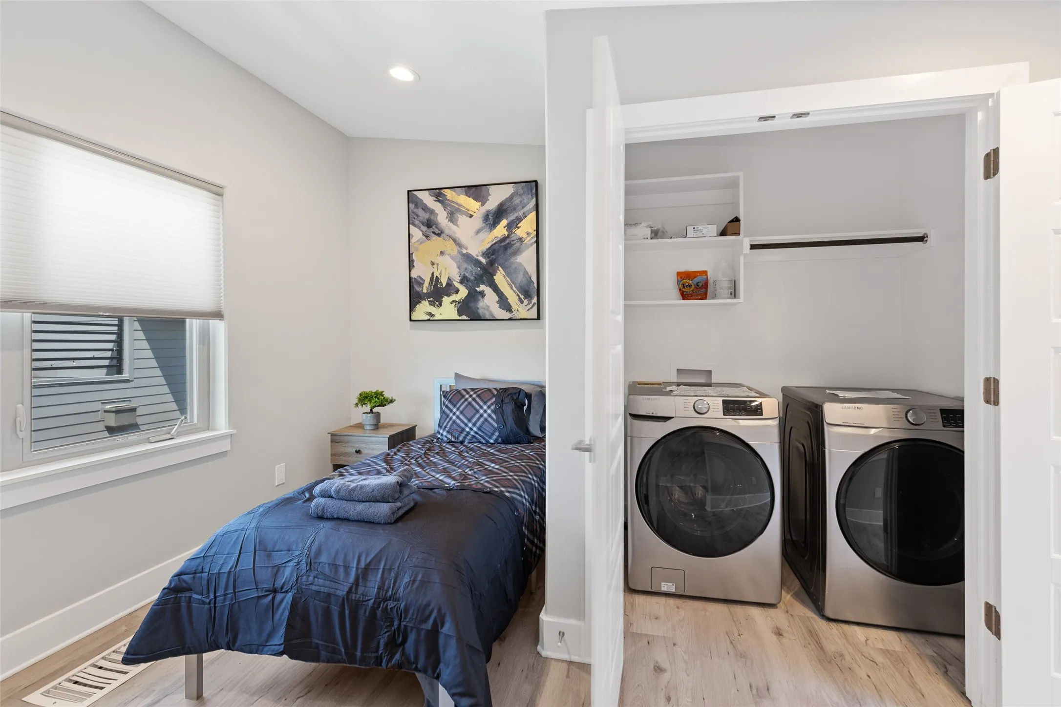 Compact bedroom with natural light from louvered window, paired with an ensuite laundry room featuring side-by-side front-load washers and dryers, wood-finish flooring throughout, and recessed ceiling lighting