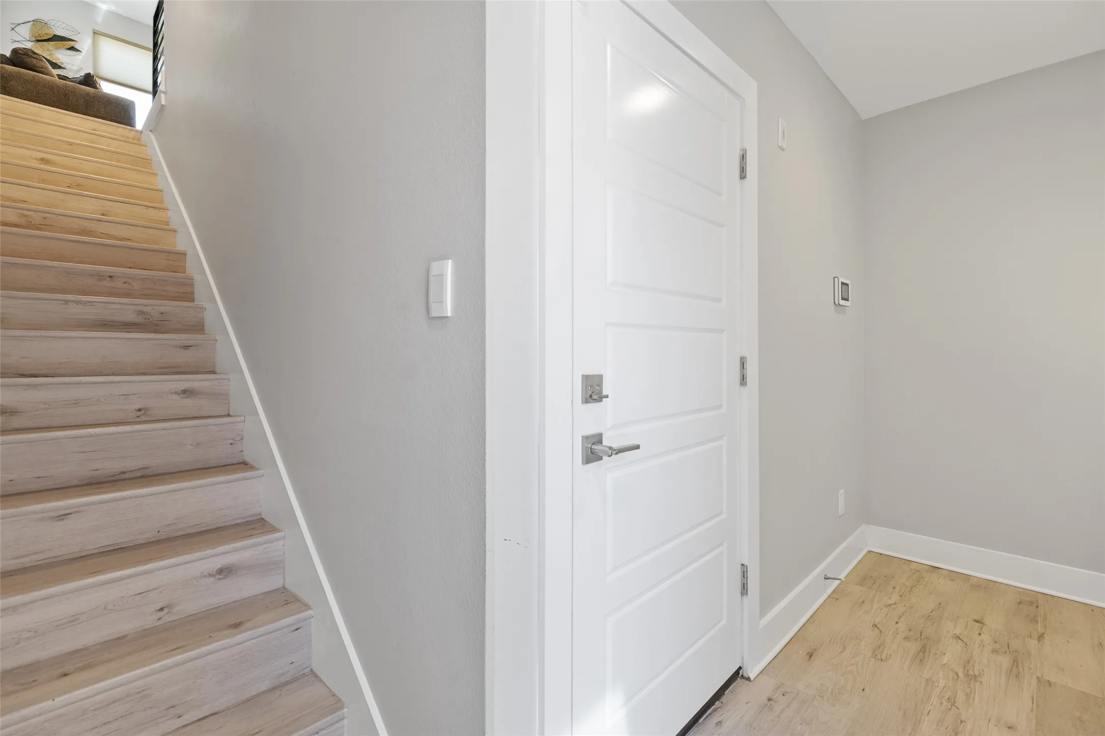 Light-toned wood-finish flooring, white panel door with brushed nickel hardware, and a staircase with wood-finish treads