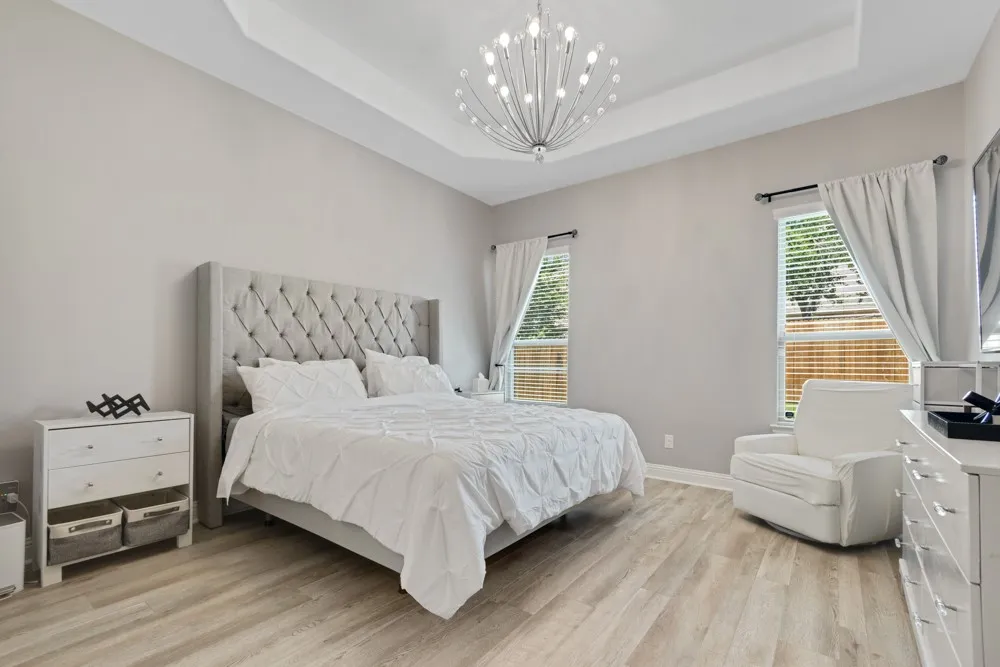 This spacious primary bedroom offers a sitting area, a tray ceiling with a contemporary chandelier, wood-finish flooring, and multiple windows with views to the backyard.