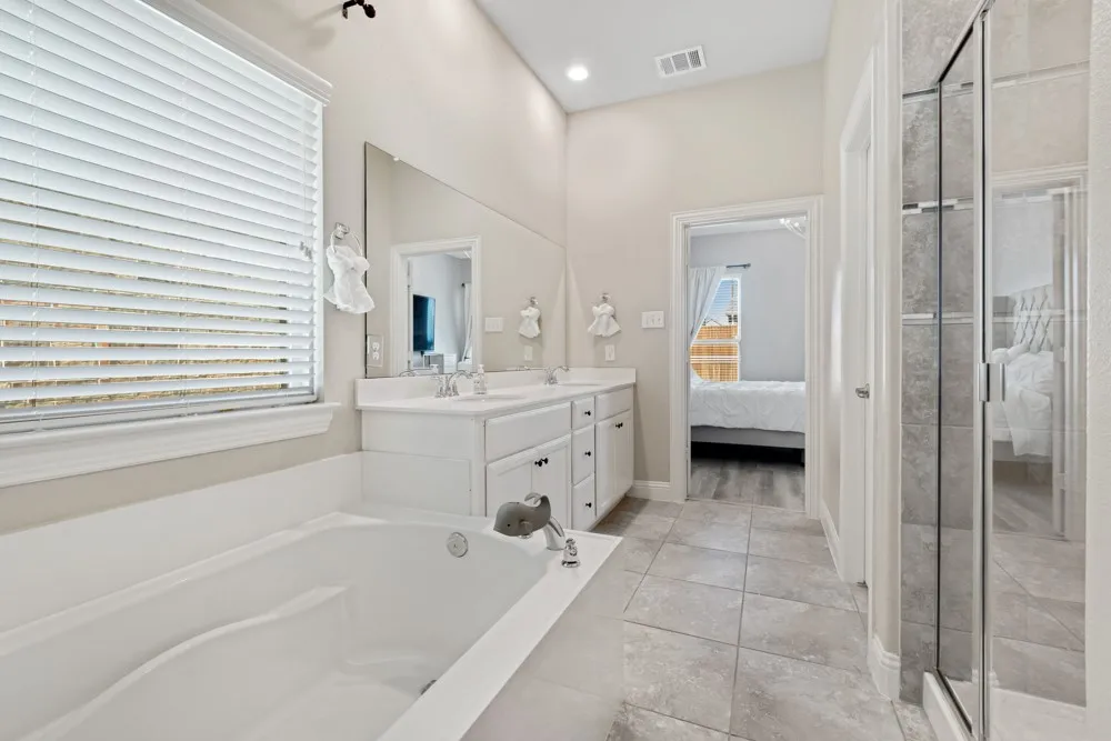 Soaking tub with adjacent window, dual sink vanity, and a glass-enclosed shower with tile surround
