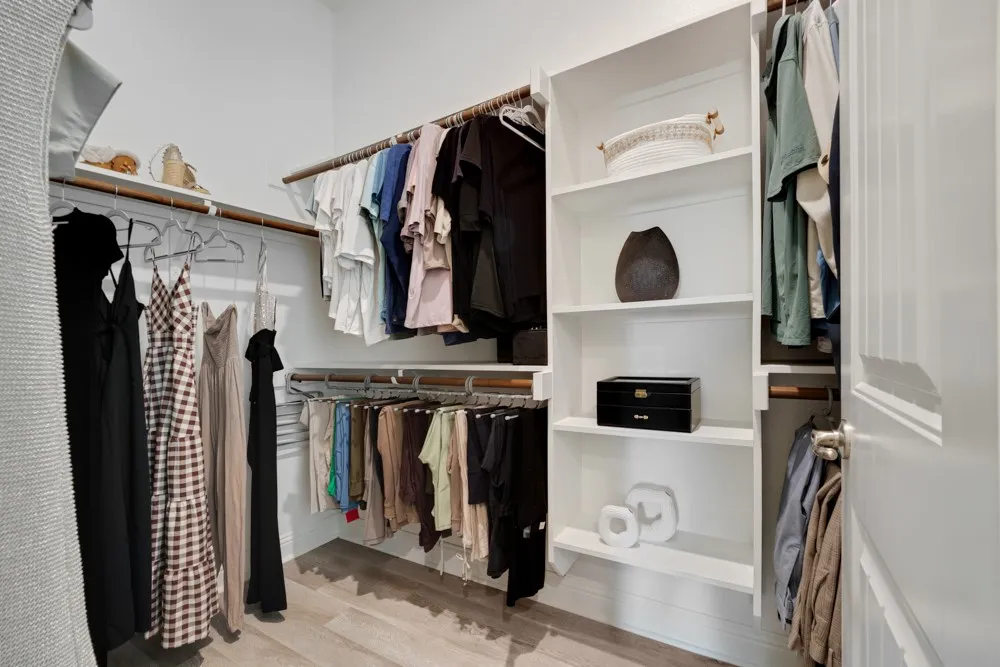 The primary closet features built-in shelving, wood-finish flooring, a white paneled door, and multiple clothing rods.  This is just a portion of the space!