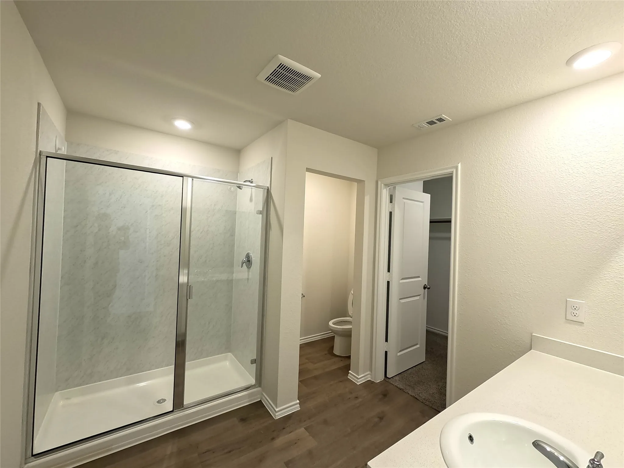 Glass-enclosed shower with a white base, white walls, wood-finish flooring, a white countertop with an integrated sink, and recessed lighting