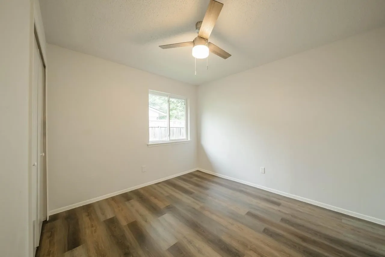 Interior room featuring wood-finish flooring, a neutral color palette, a ceiling fan with integrated lighting, and a window with a slider mechanism