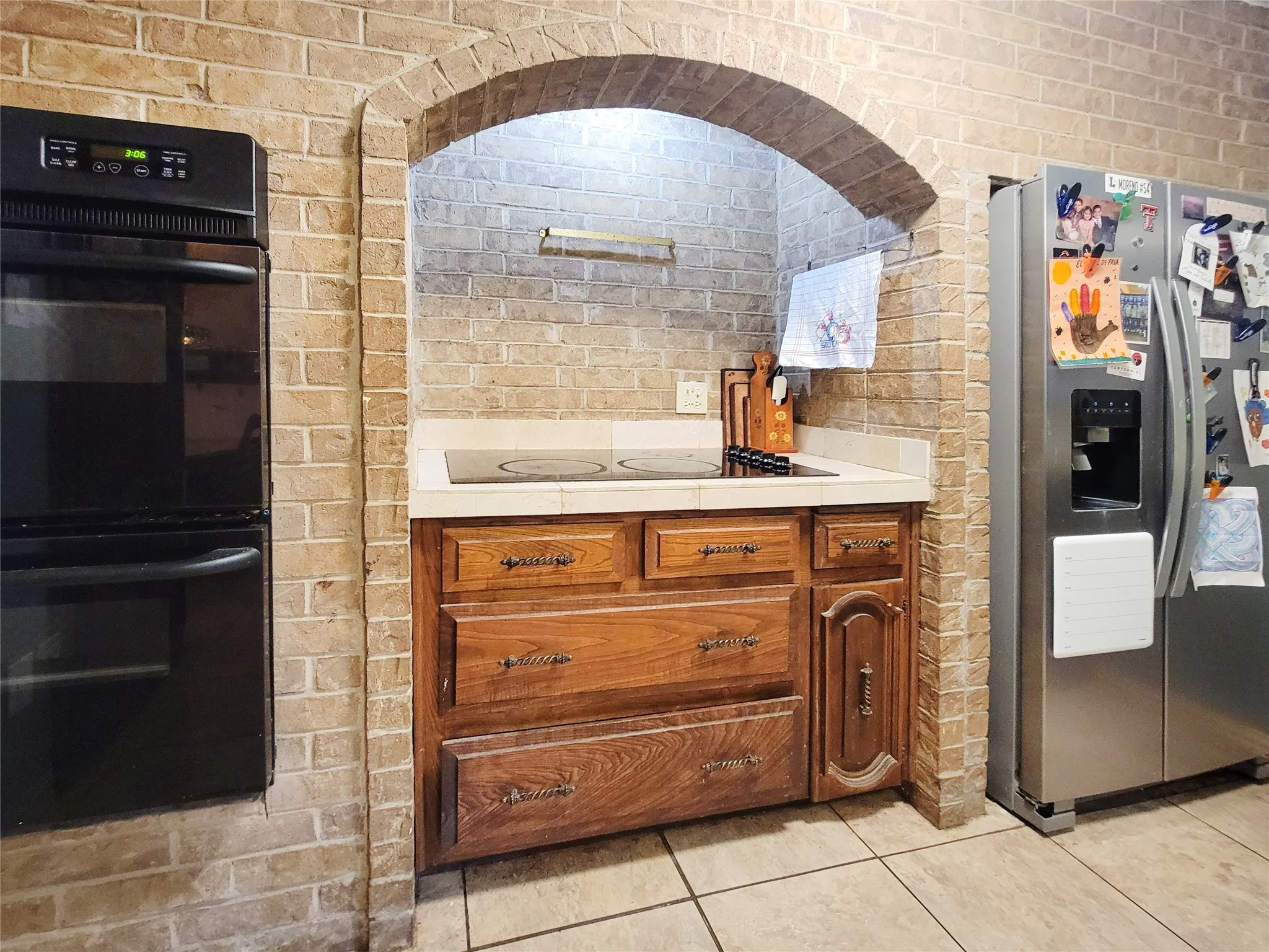 Brick-finish walls with an arched alcove, built-in double ovens, light-toned countertop, integrated cooktop, and wood-finish cabinetry