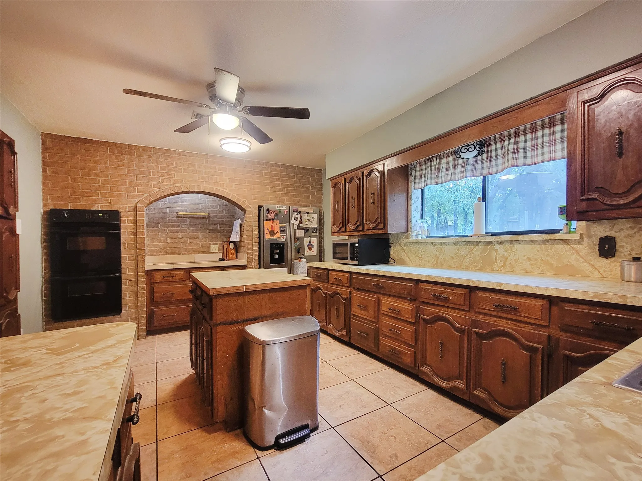 Brick accent wall with arched opening, double wall oven, wood-finish cabinetry, tile flooring, and a ceiling fan