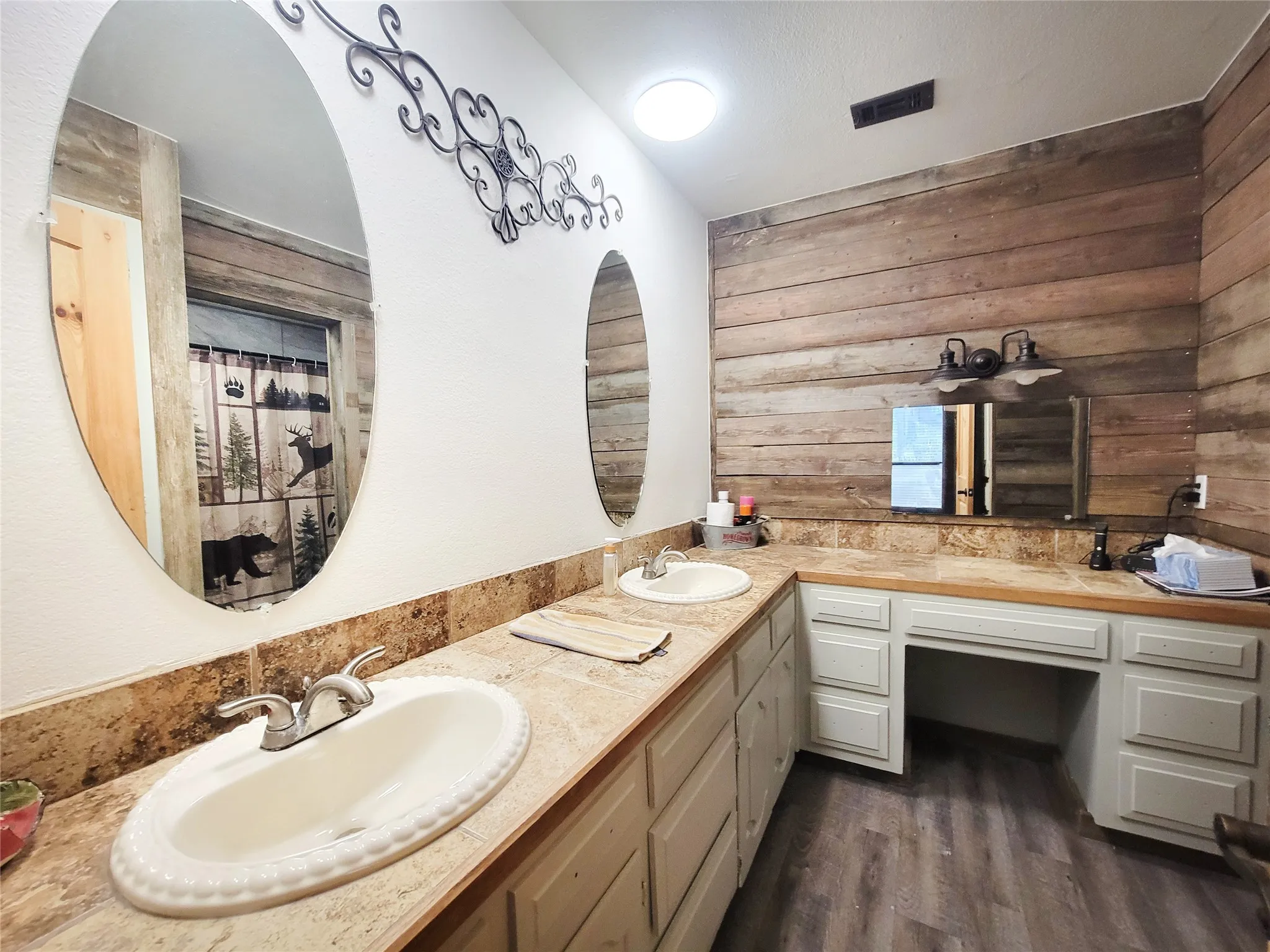 Dual oval vanity mirrors, wood-finish accent wall, built-in cabinetry, dual basins, and wood-finish flooring