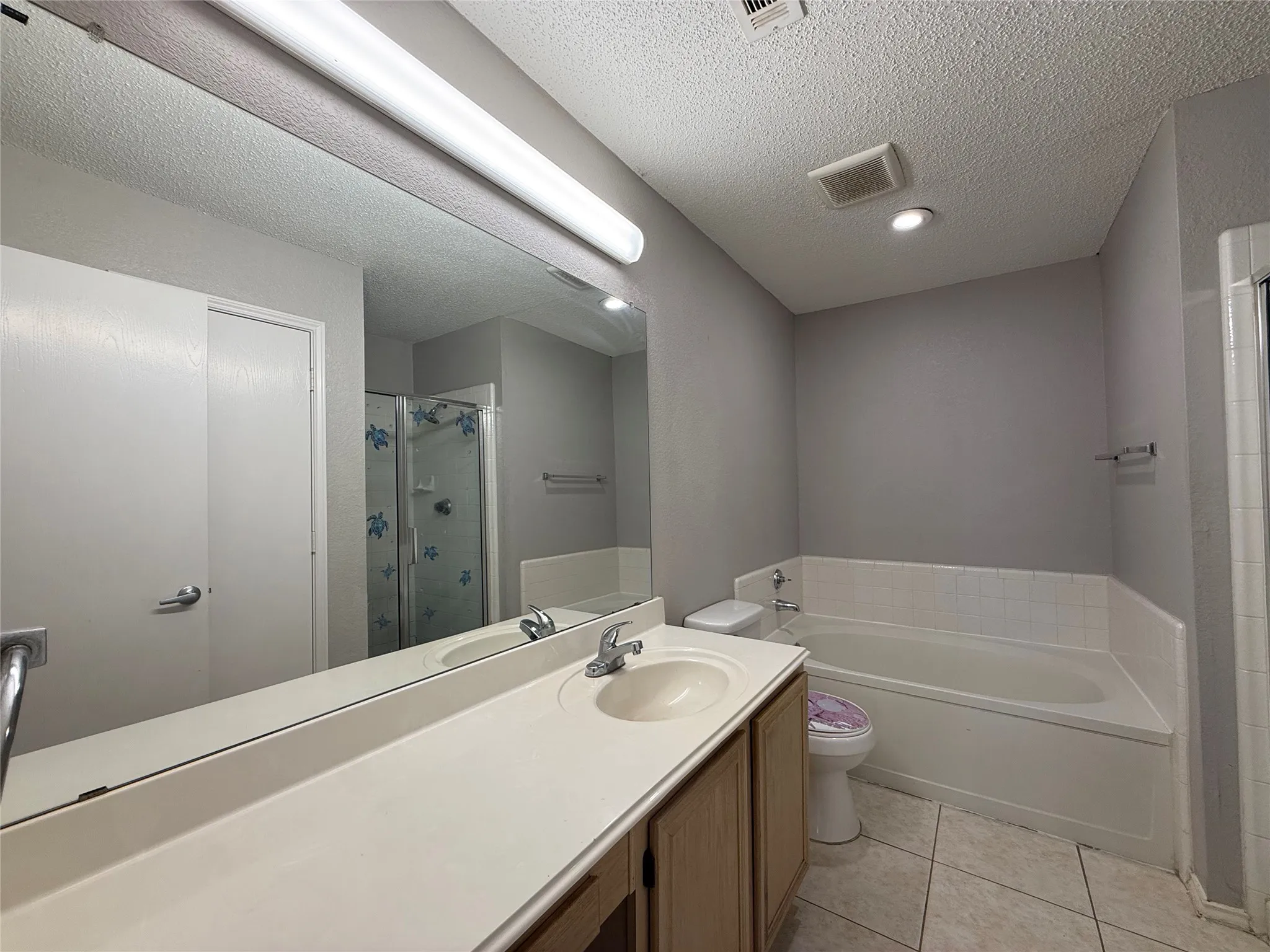 Full-width wall mirror, wood-finish vanity cabinet with a neutral integrated countertop and sink, soaking tub with a tiled surround, and ceramic tile flooring