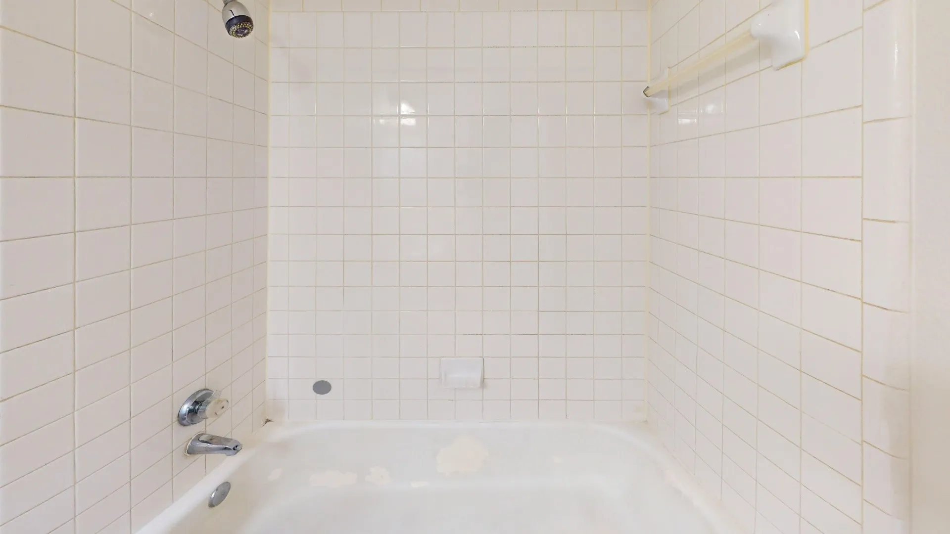 Bathtub enclosure featuring white square tile surround, wall-mounted shower head, metal faucet hardware, and integrated soap dish