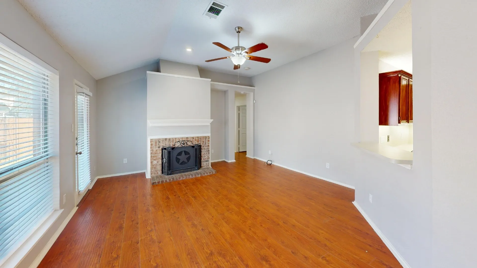 Vaulted ceiling living area featuring wood-finish flooring, a brick fireplace with a mantle, a ceiling fan, and a pass-through window to the kitchen