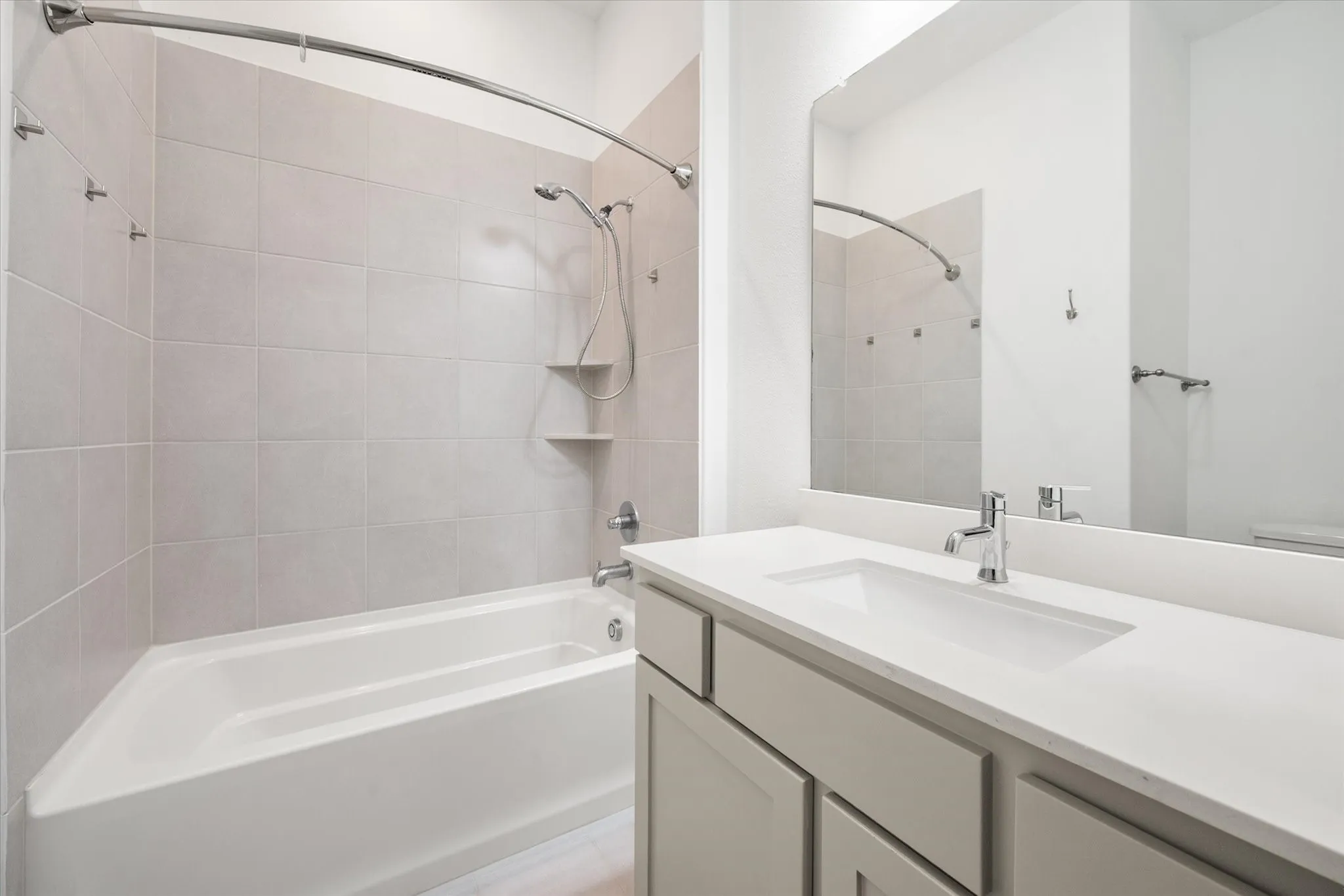 White rectangular undermount sink, chrome faucet, light grey vanity with panel doors, white countertop, and tiled tub surround with shower fixture