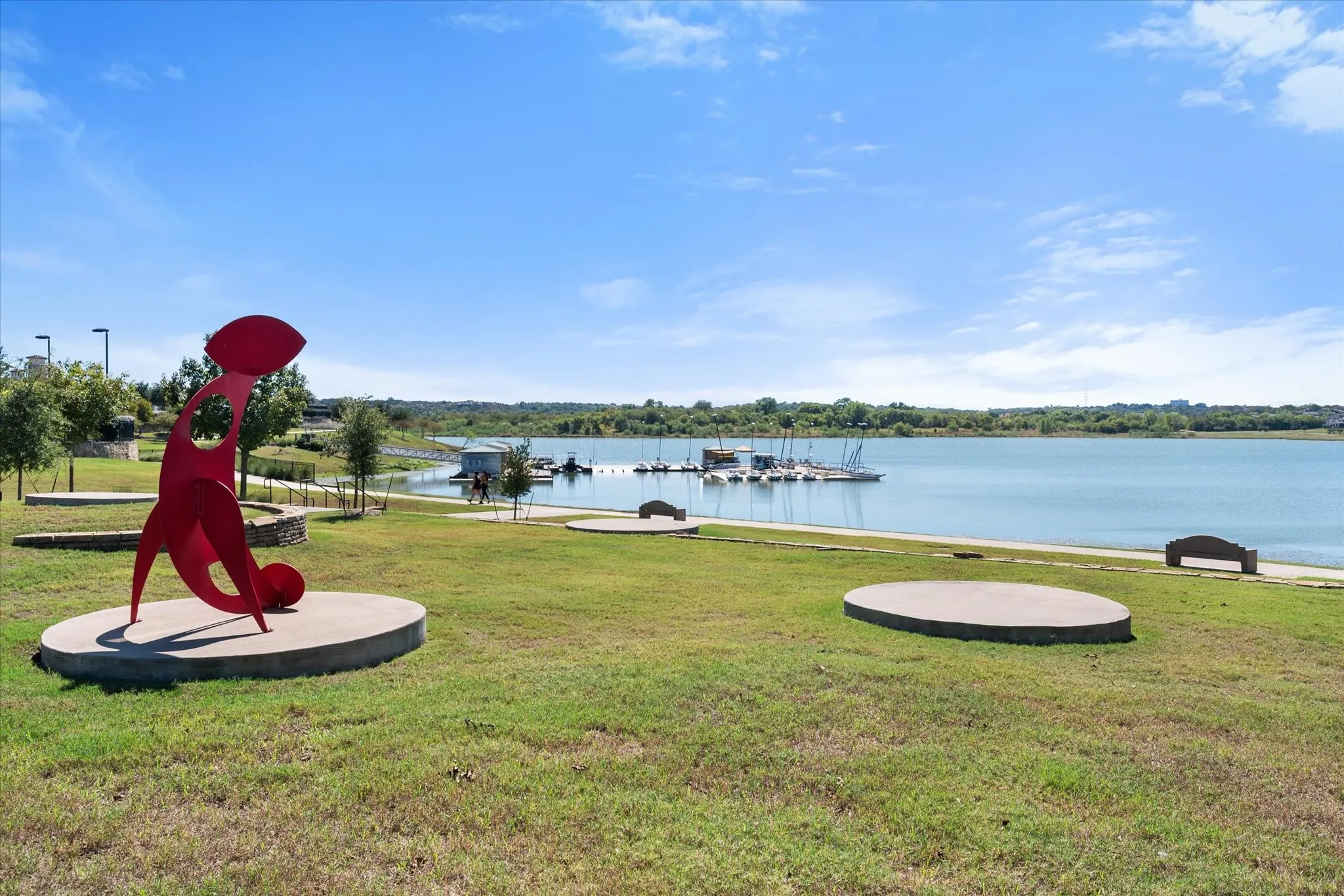 Expansive green space featuring a large body of water, a dock with moored boats, and a concrete pathway