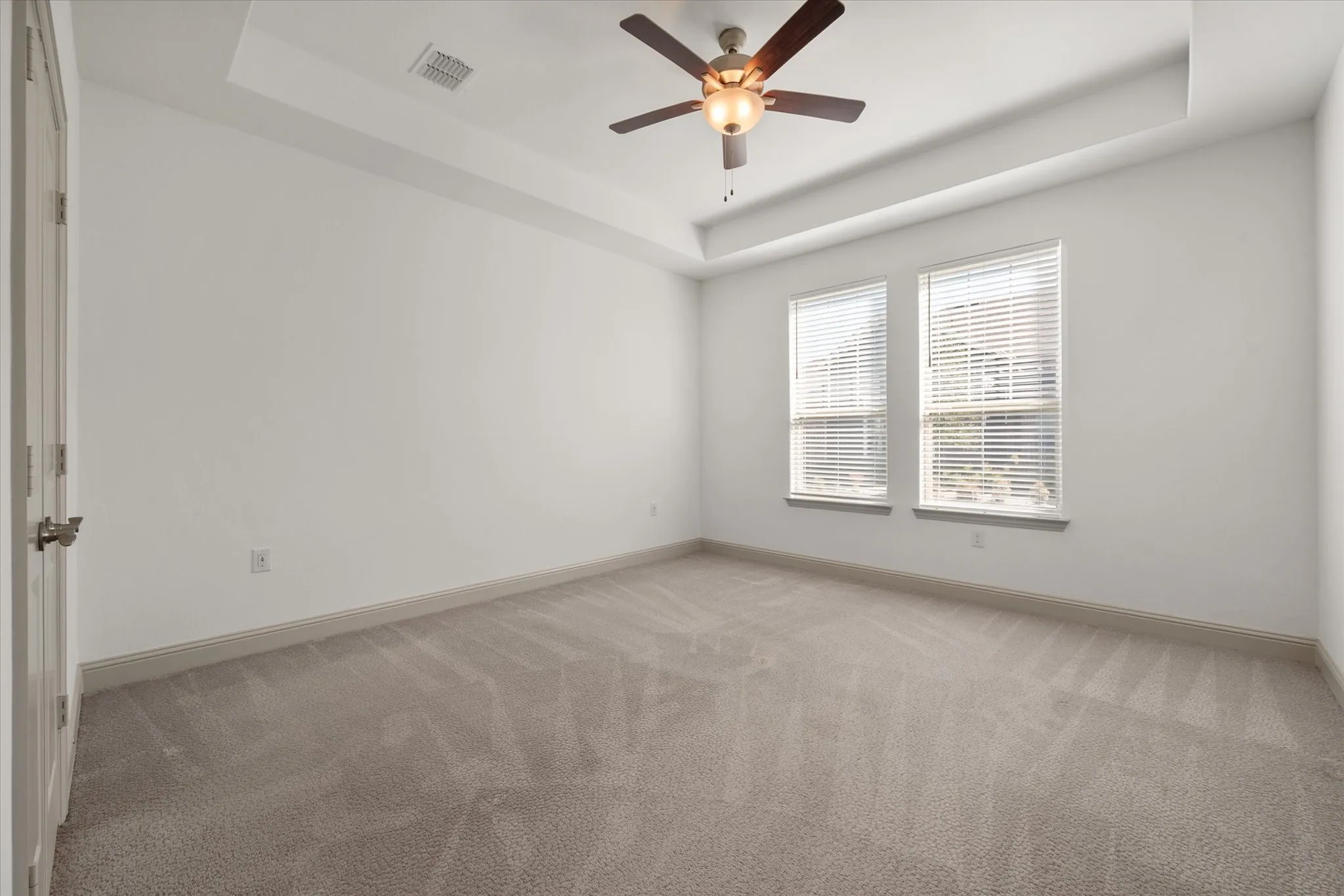 Tray ceiling with a ceiling fan, two windows with blinds, and neutral-toned carpeting