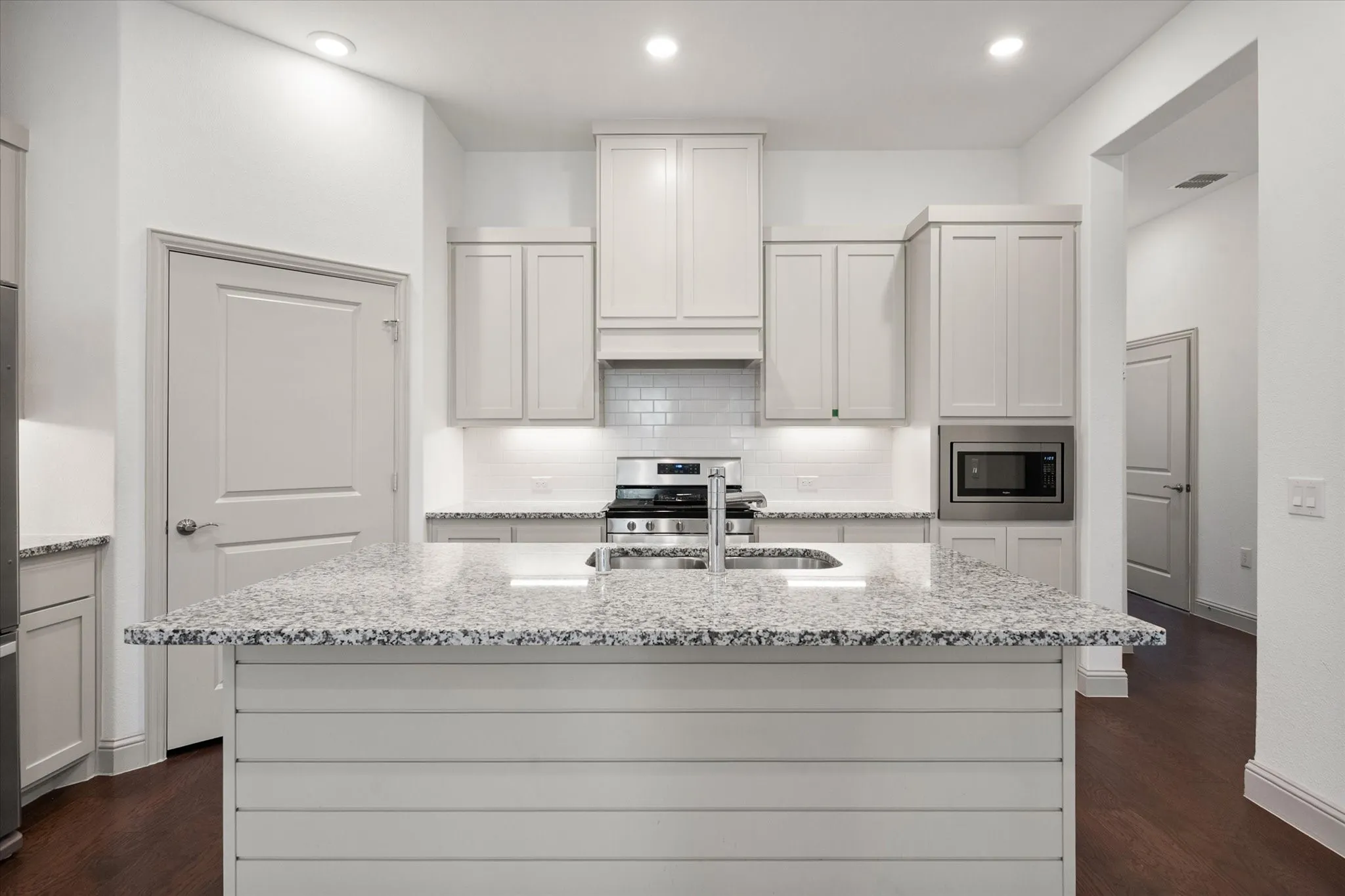 Kitchen featuring a central island with a built-in sink, stainless steel appliances, light cabinetry, stone countertops, and a subway tile backsplash