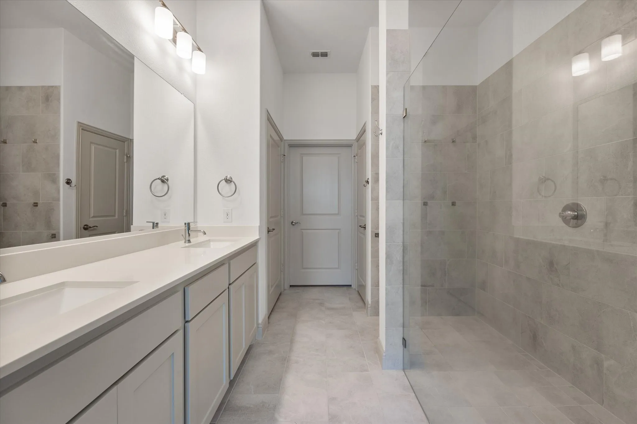 Dual vanity with integrated sinks, expansive mirror, contemporary light fixtures, glass-enclosed shower, and large format tile flooring