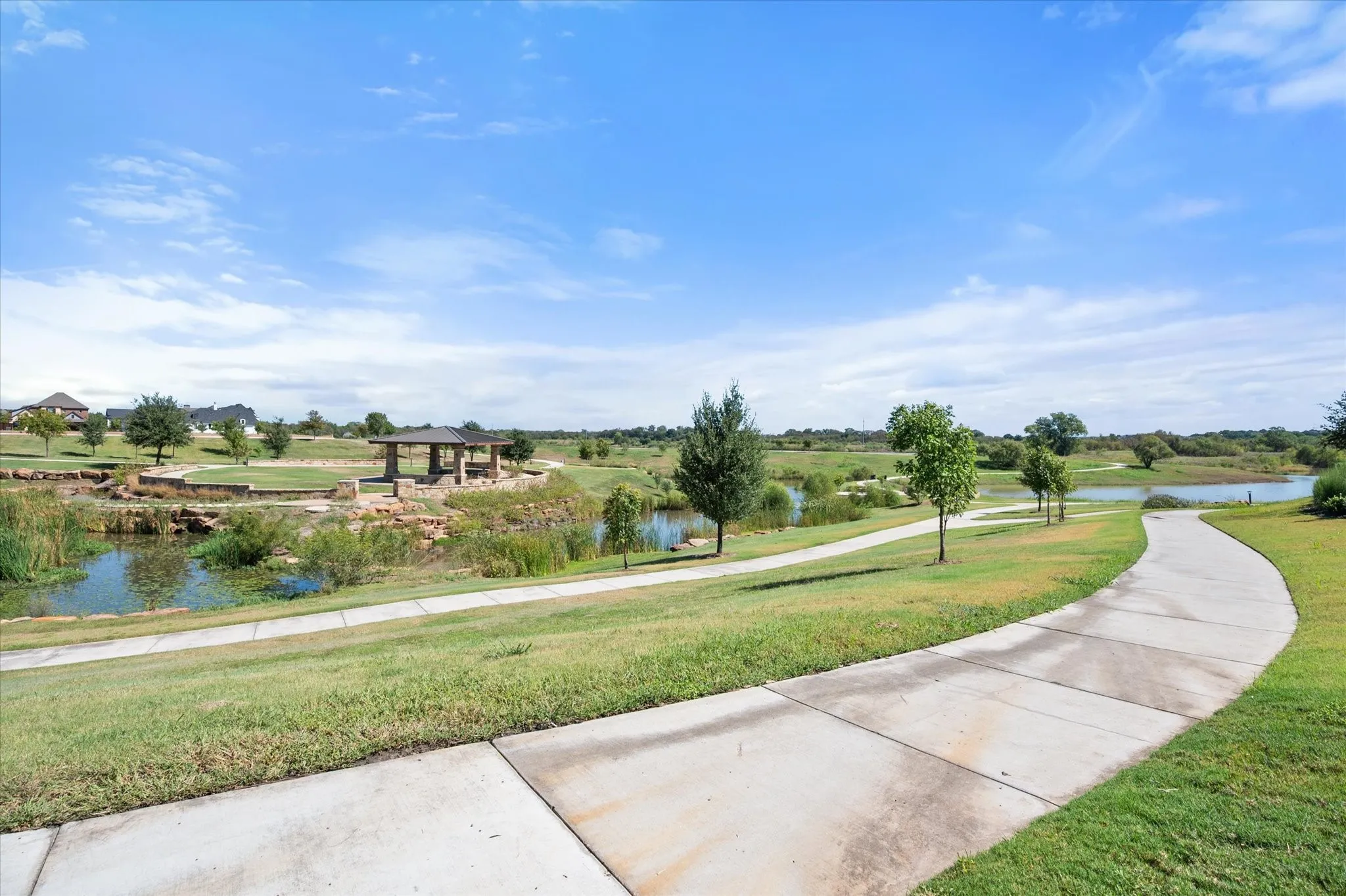 Paved walking path bordered by expansive green space, mature trees, and a water feature