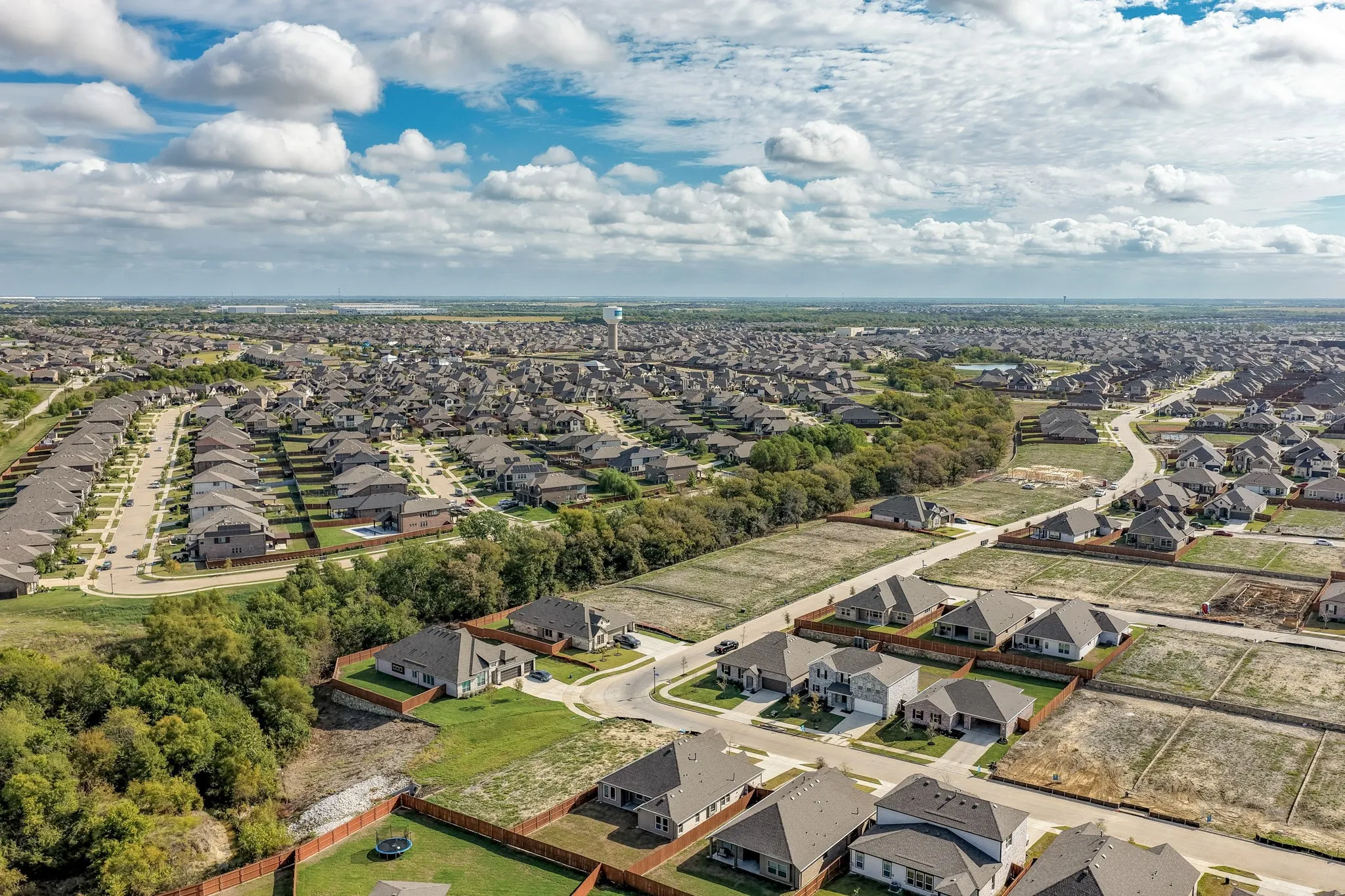 Aerial perspective of a residential community featuring winding paved streets, single-story homes with composite shingled roofs, wood privacy fencing, and surrounding green space