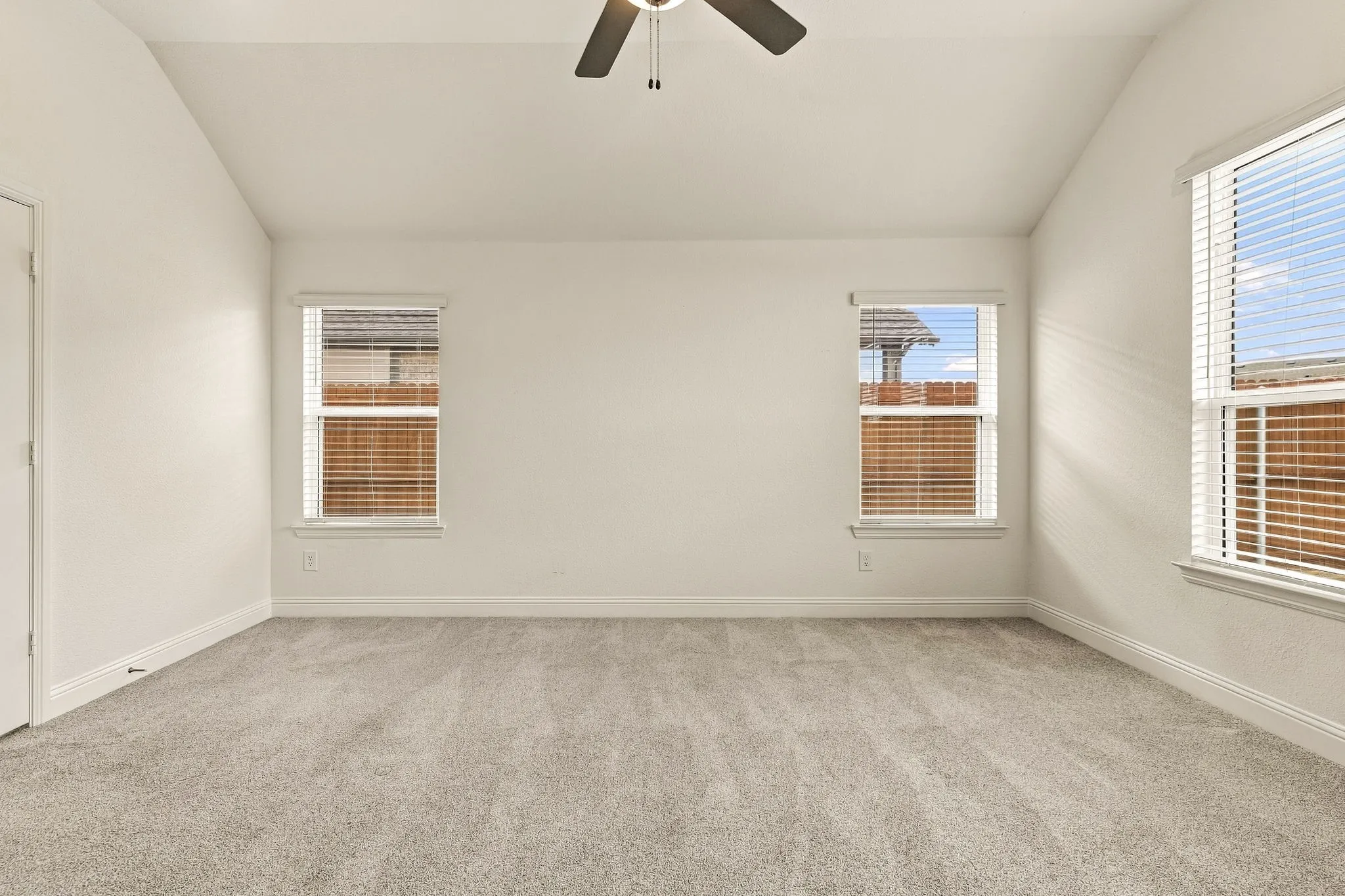 Vaulted ceiling with a centrally mounted ceiling fan, neutral wall-to-wall carpeting, and multiple windows with installed horizontal blinds
