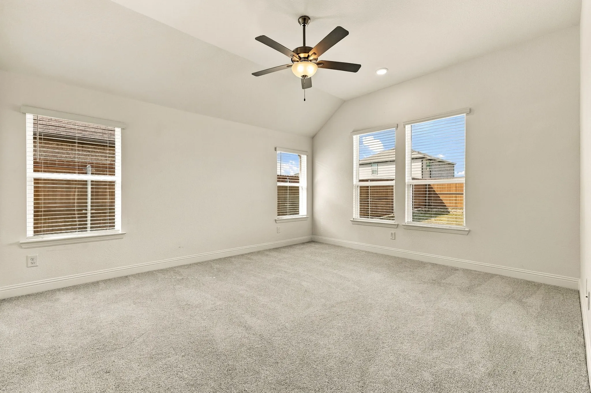 Vaulted ceiling featuring a central five-blade ceiling fan with integrated lighting, neutral wall-to-wall carpeting, and multiple windows with horizontal blinds