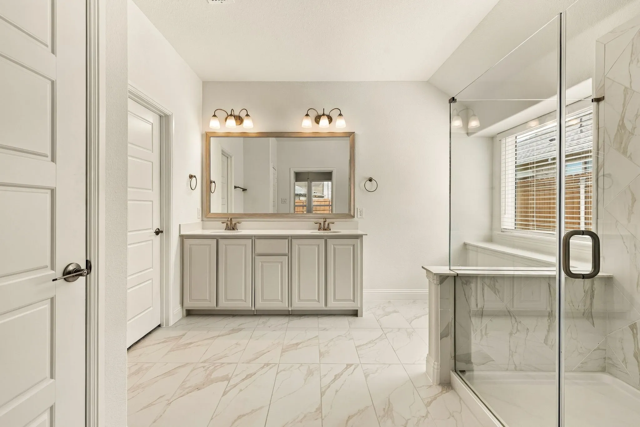 Dual vanity with light-toned cabinetry and solid surface countertop, expansive framed vanity mirror, glass-enclosed shower with stone-finish tile surround, and neutral stone-finish floor tiling
