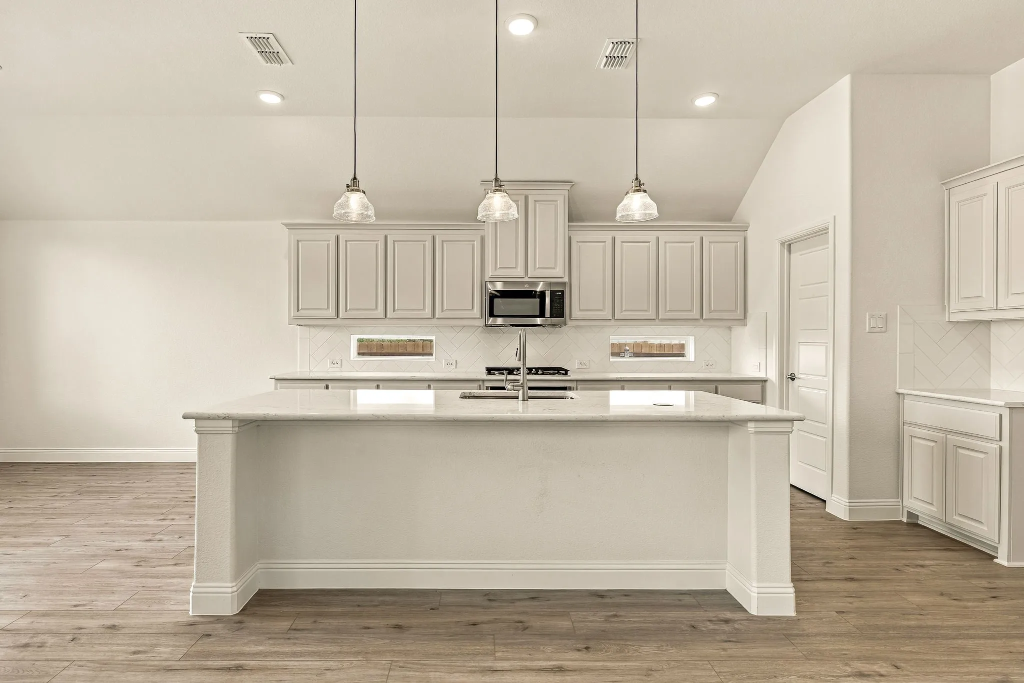 Kitchen featuring a central island with stone countertop, wood-finish flooring, white shaker-style cabinetry, a herringbone-patterned backsplash, and trio of pendant lights