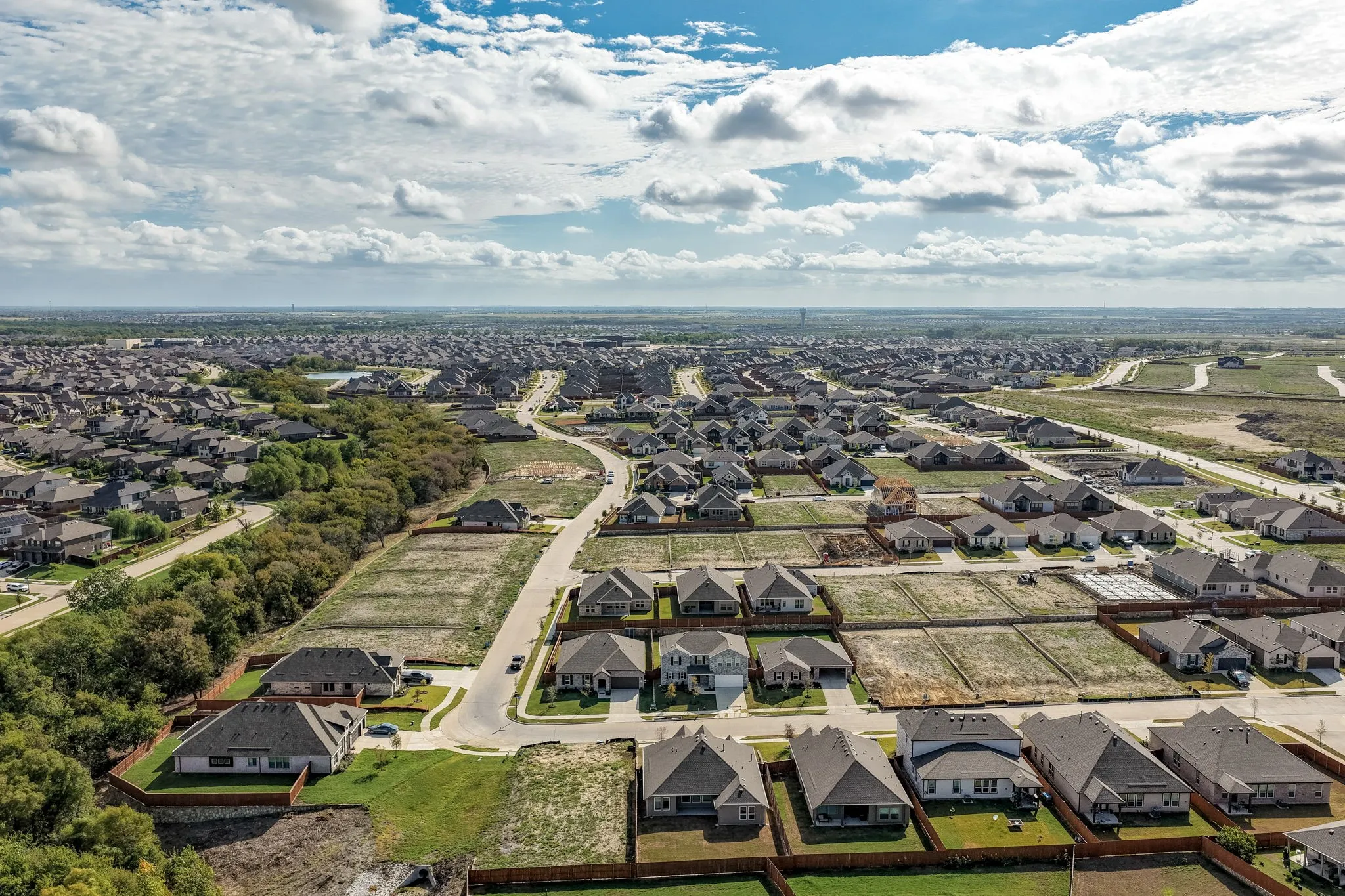 Aerial perspective of a planned residential development featuring single-family residences, asphalt paved streets, wood-fencing property boundaries, and mature perimeter trees