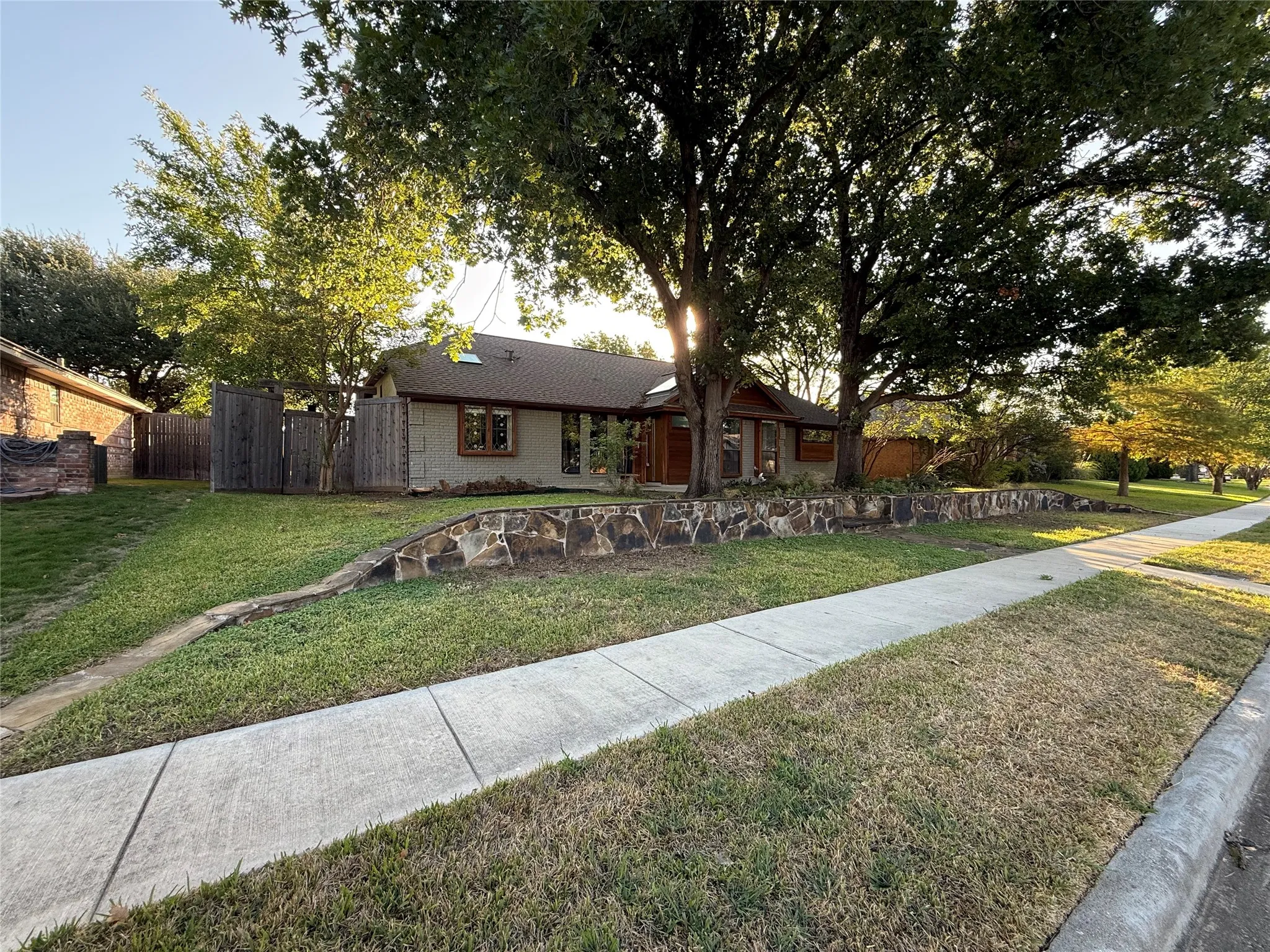 Single-story residence featuring a brick facade, multi-gabled roofline, stone retaining wall, and mature shade trees