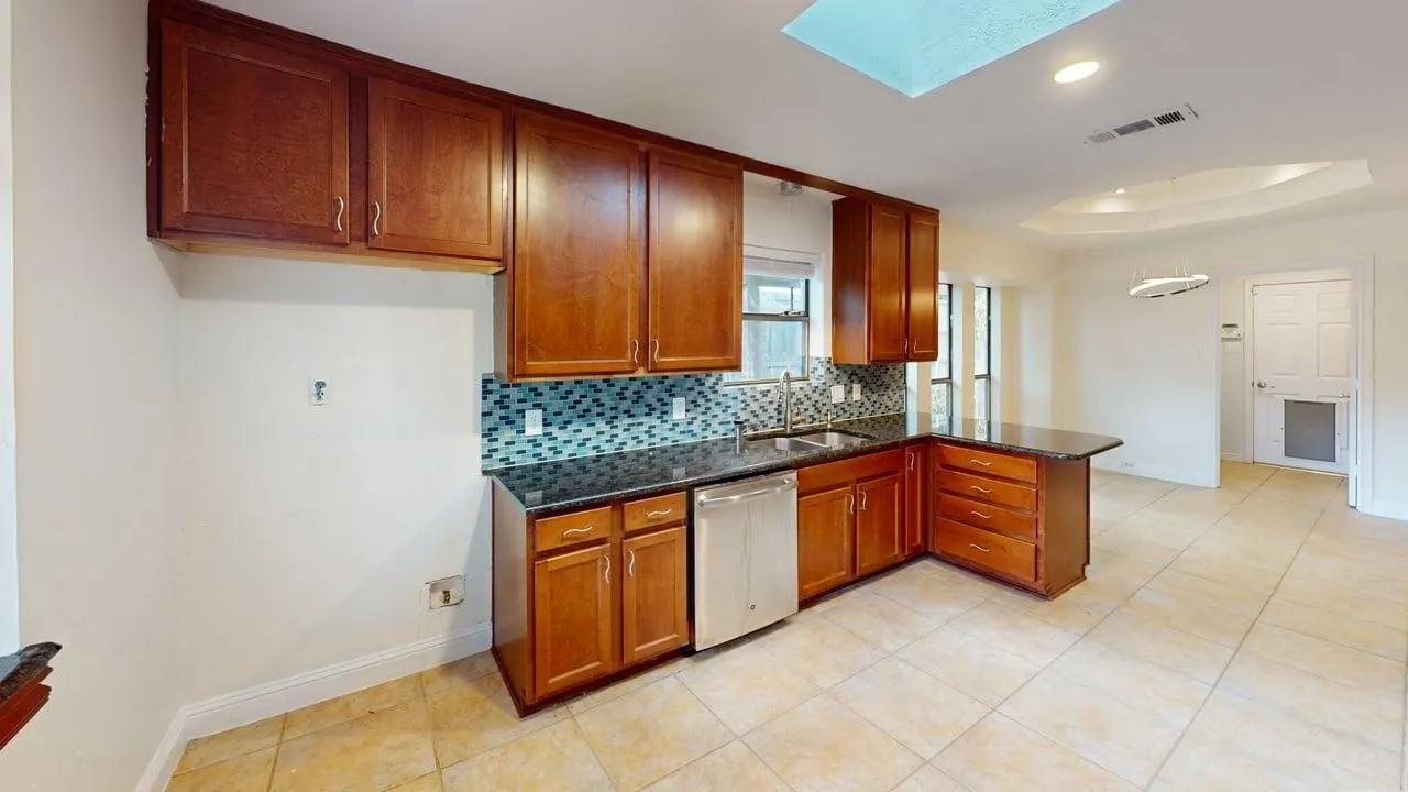 Kitchen featuring wood cabinetry, dark stone-finish countertops, a mosaic tile backsplash, a stainless steel dishwasher, and a ceiling skylight