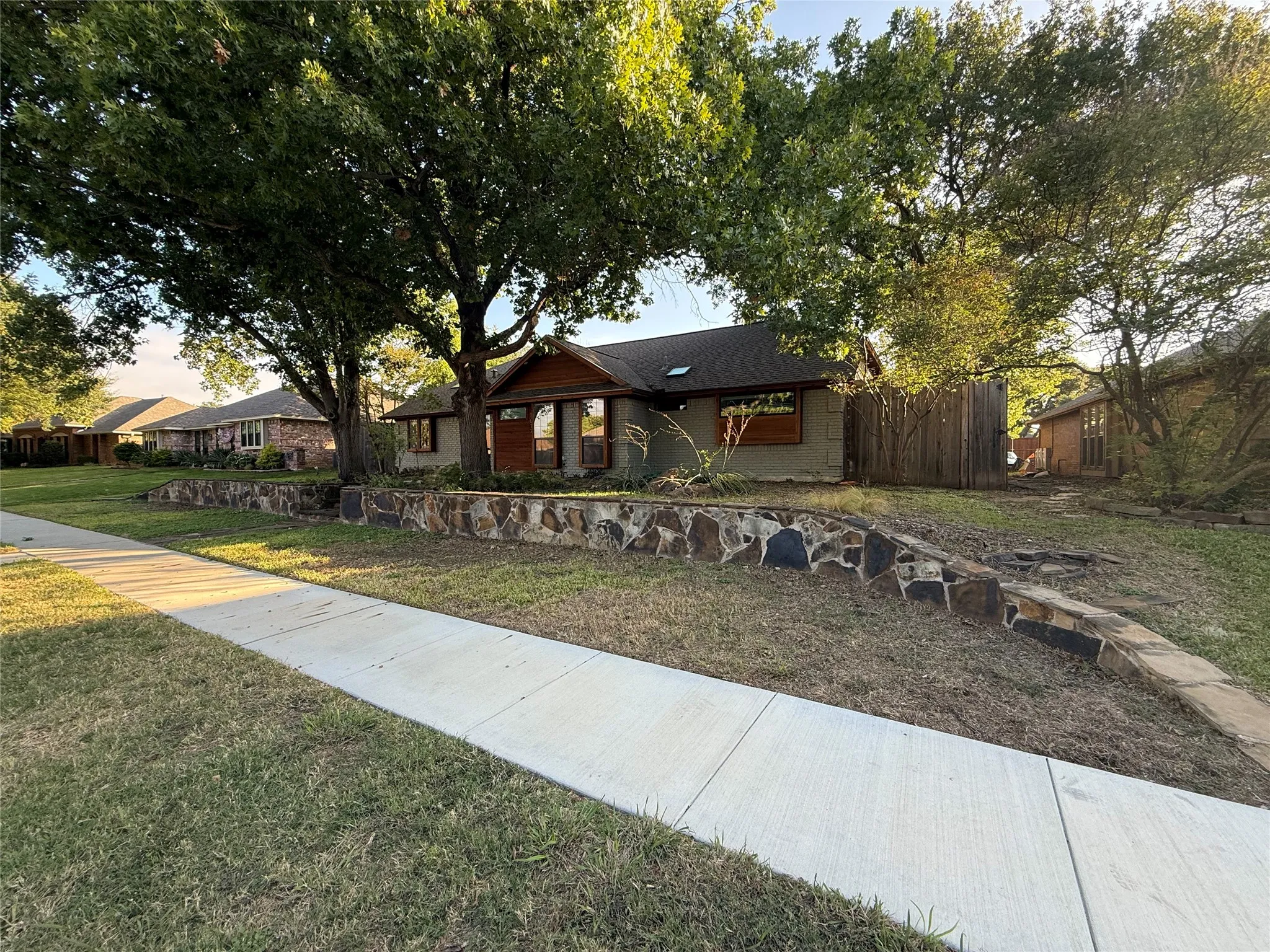 Exterior elevation featuring a stone-clad retaining wall, gabled roofline, skylight, and mature shade trees