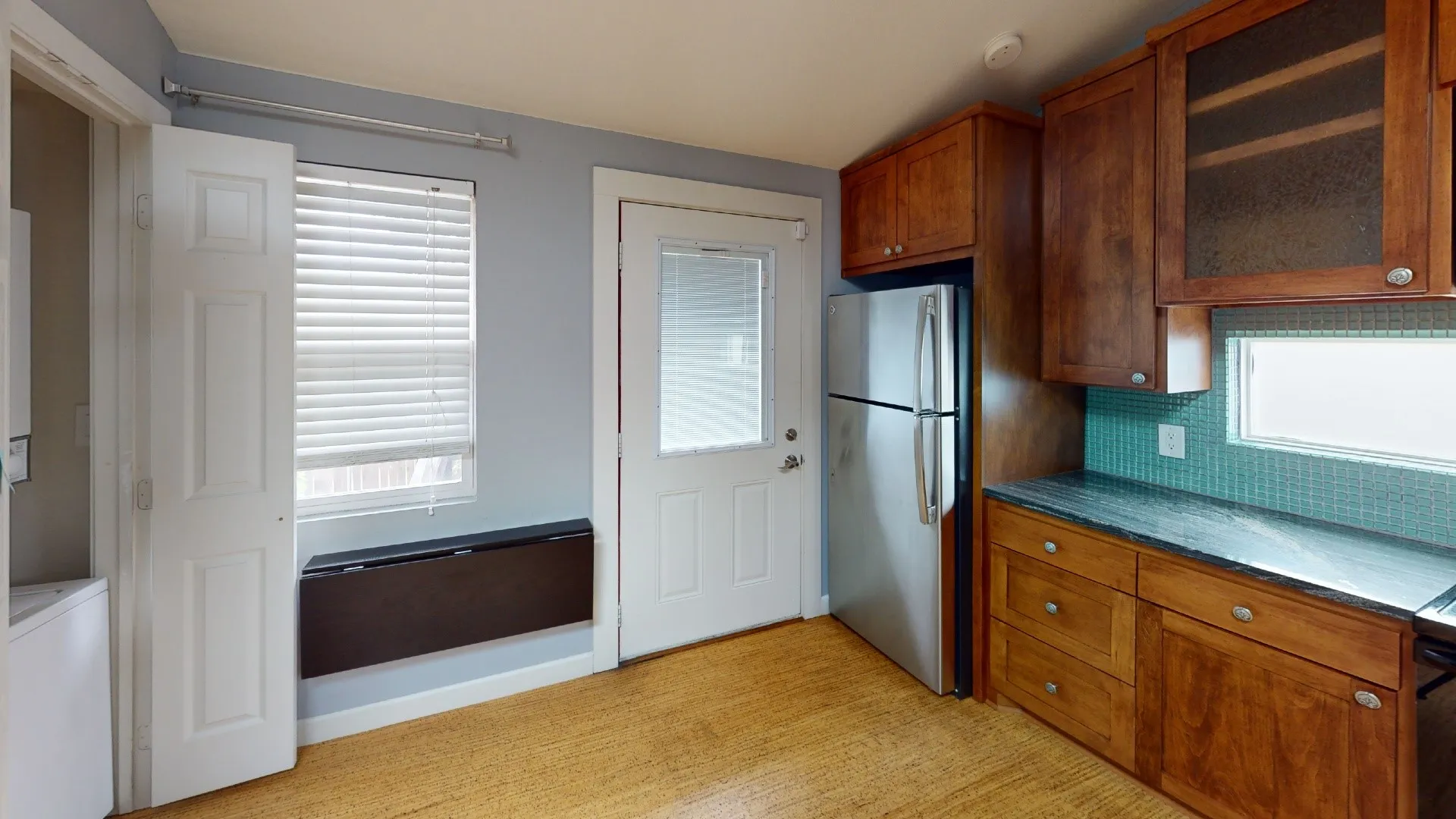 Kitchen featuring wood cabinetry, dark stone-finish countertops, a teal tile backsplash, and cork flooring